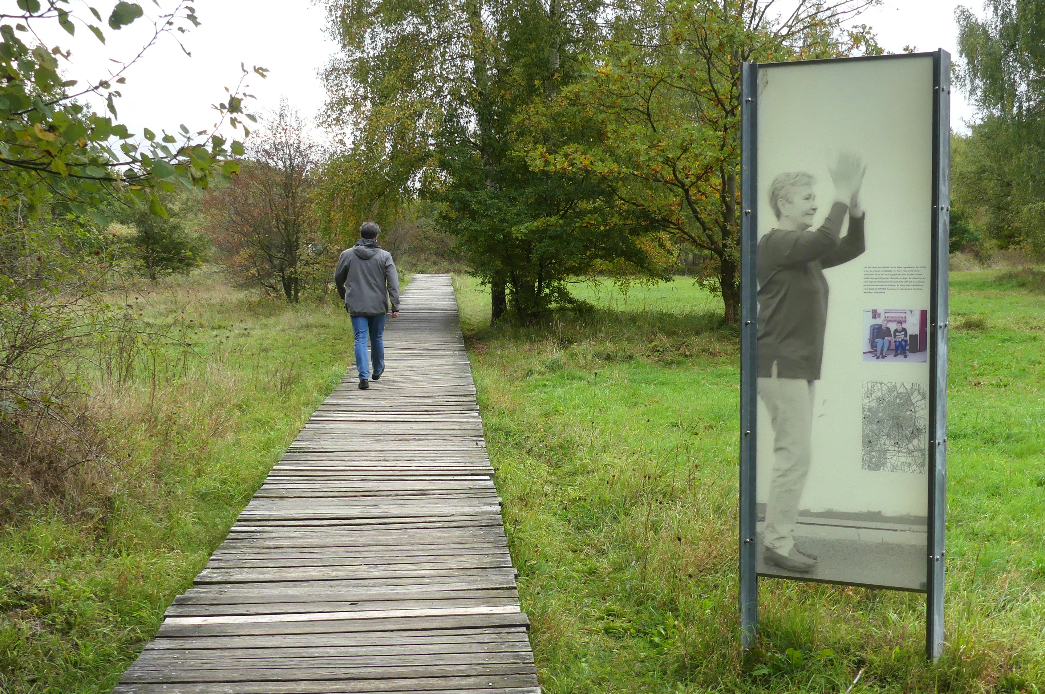 A person walks along a wooden boardwalk through a grassy forest clearing, passing a tall rectangular sign displaying a black and white photographic portrait.