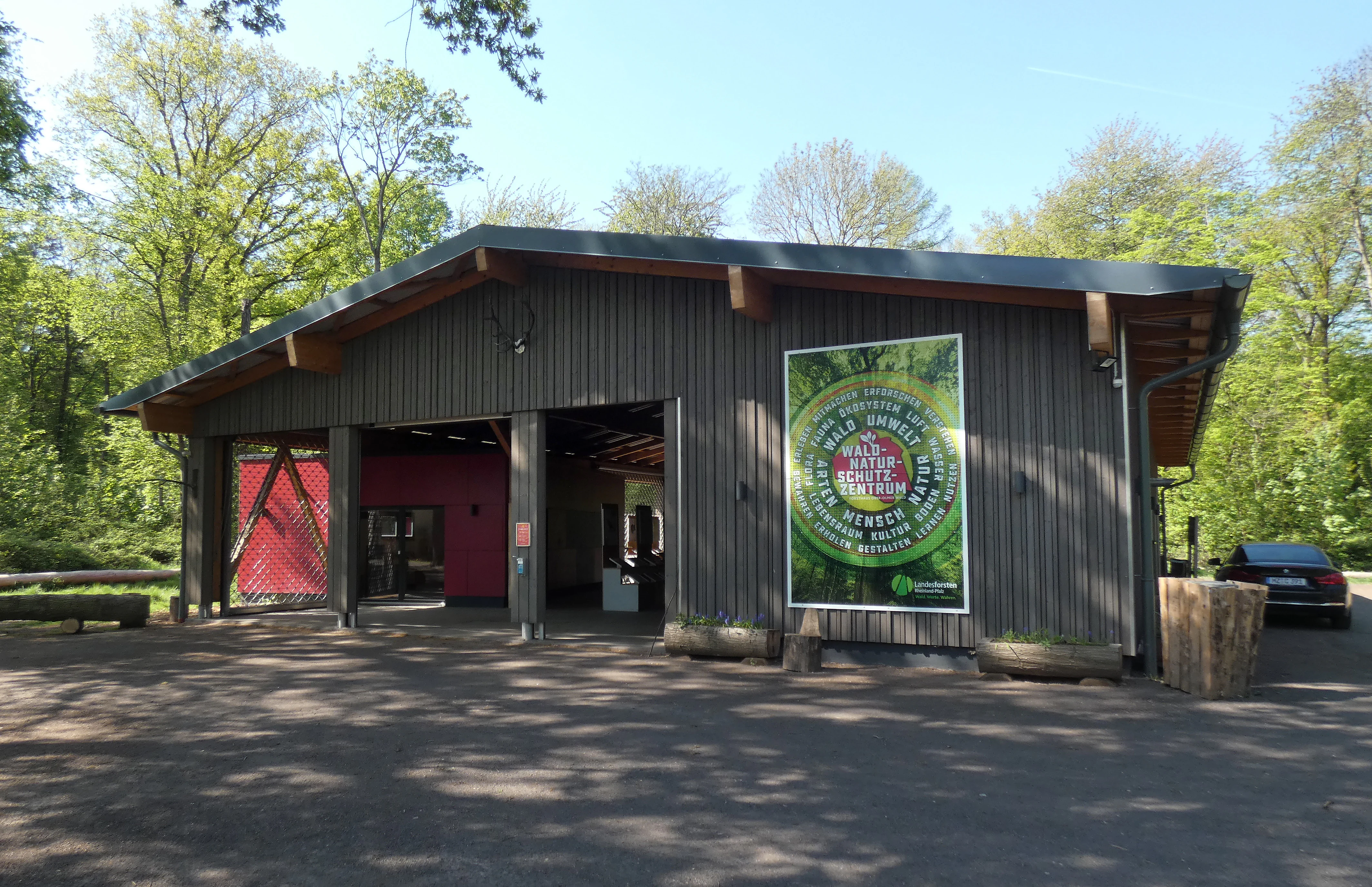 A modern, grey-paneled single-story building with a wide, open-air entrance hall, situated in a forest clearing at the Ober-Olmer Wald.