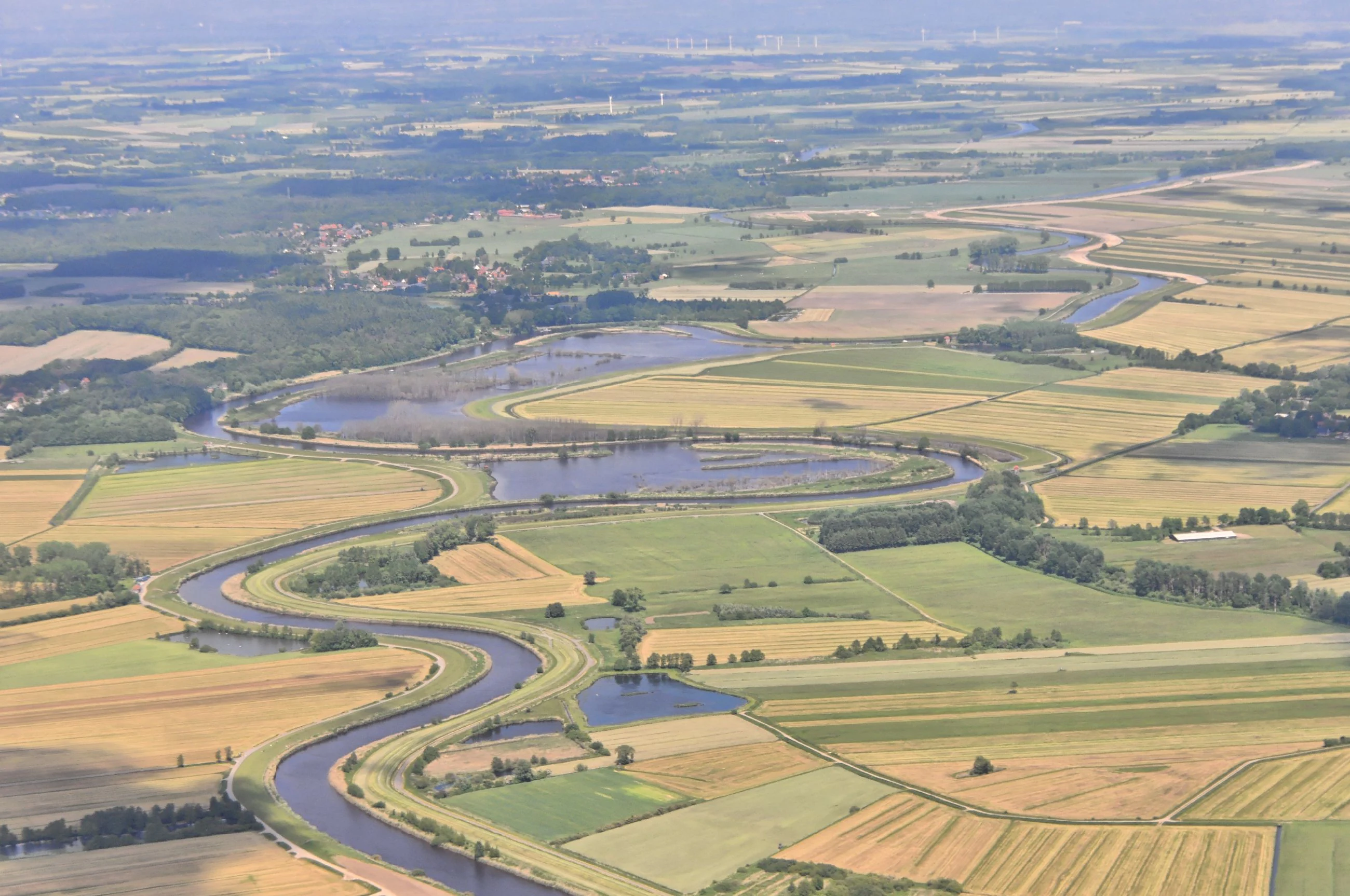 An aerial view of a winding river flowing through a mosaic of agricultural fields and wetland areas, characterized by green and golden patches of vegetation.