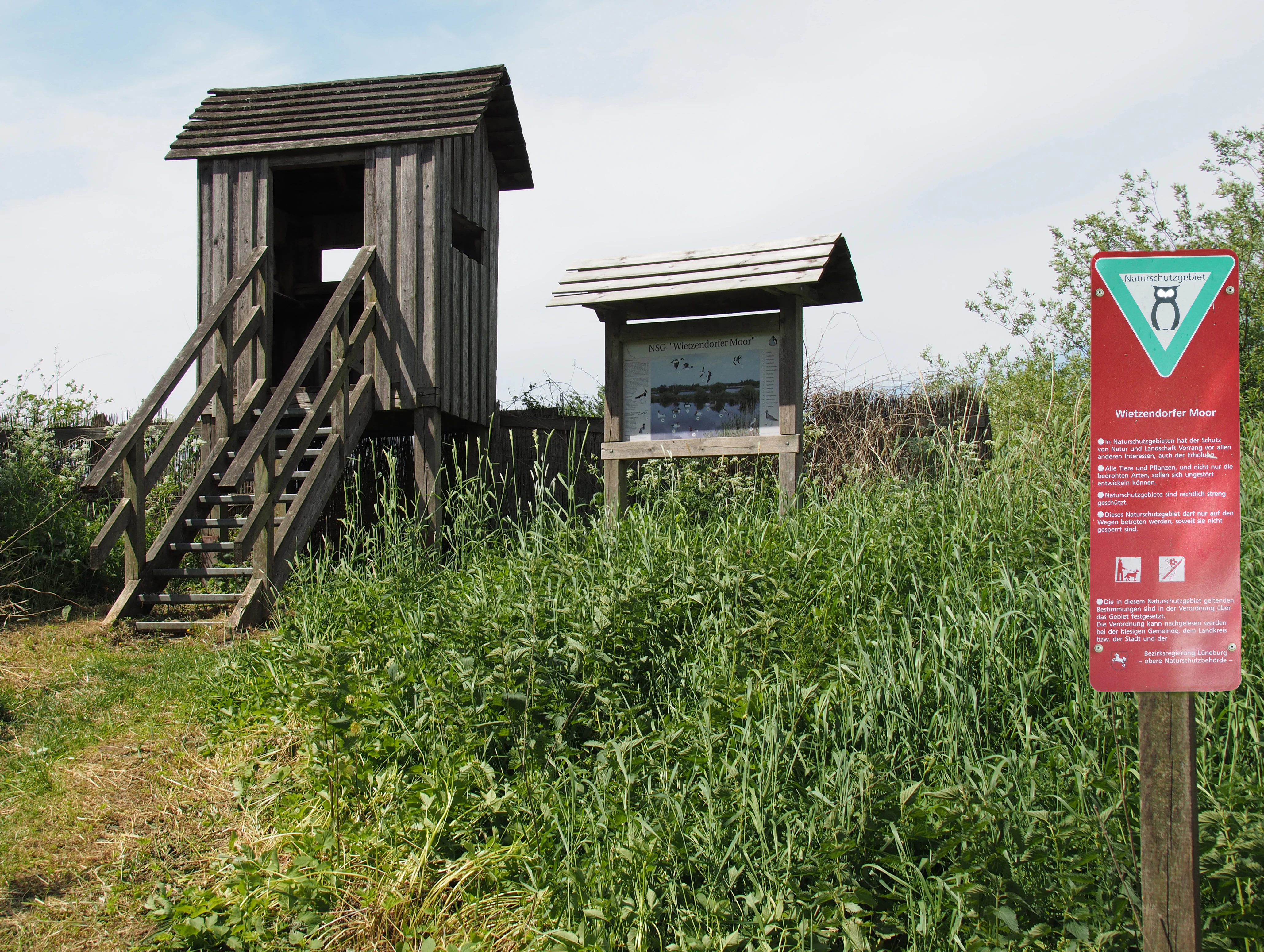 A small wooden observation tower with stairs sits in a grassy, partially overgrown field next to an information display and a red nature reserve sign with an owl logo.
