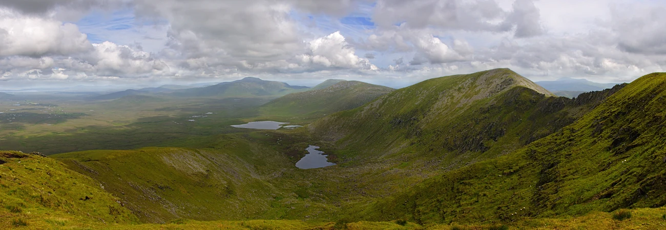 Panoramic view of rolling green mountains, valleys, and lakes under a partly cloudy sky with blanket bog in the foreground