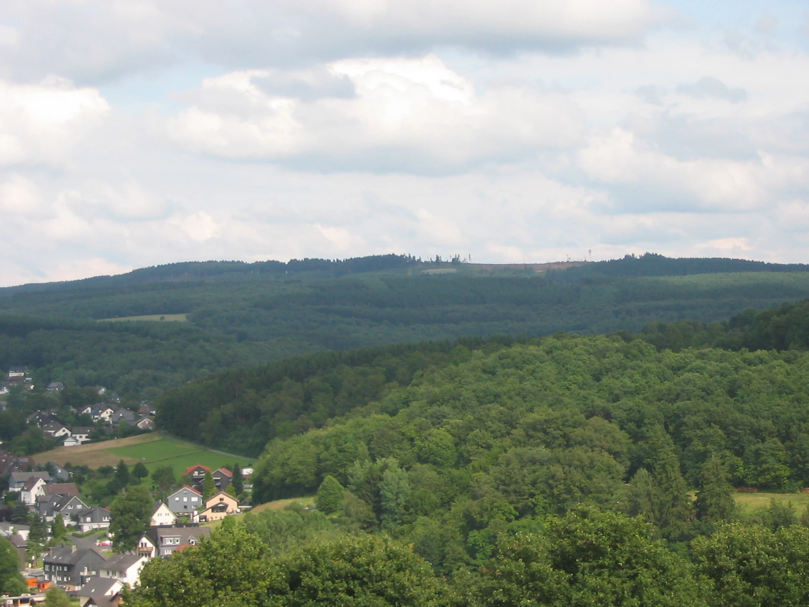 A wide view of a forested hill ridge under a cloudy sky, with a small village nestled in the valley in the foreground.