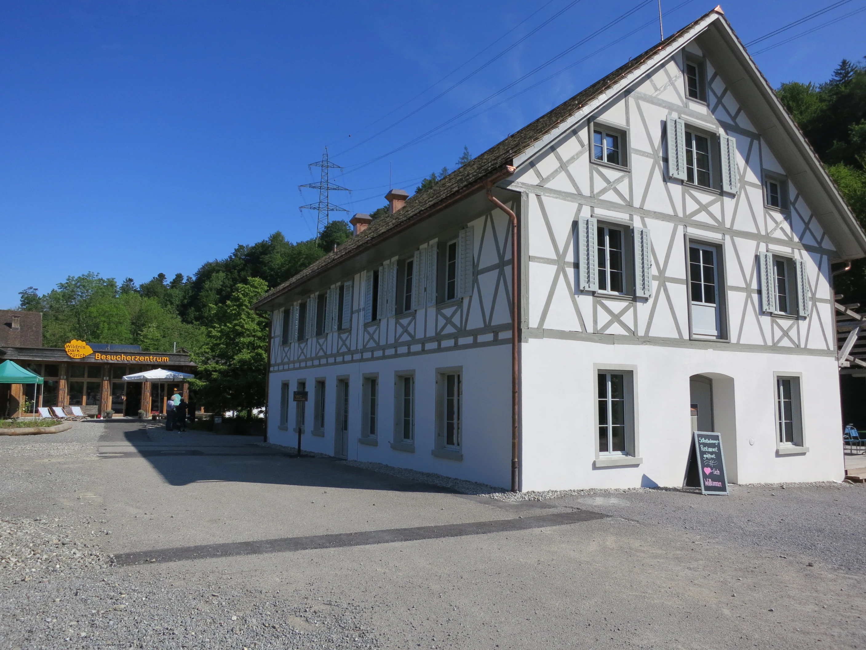 A traditional timber-framed building serving as a visitor center, situated in a park area with a gravel courtyard and forested hillside in the background.