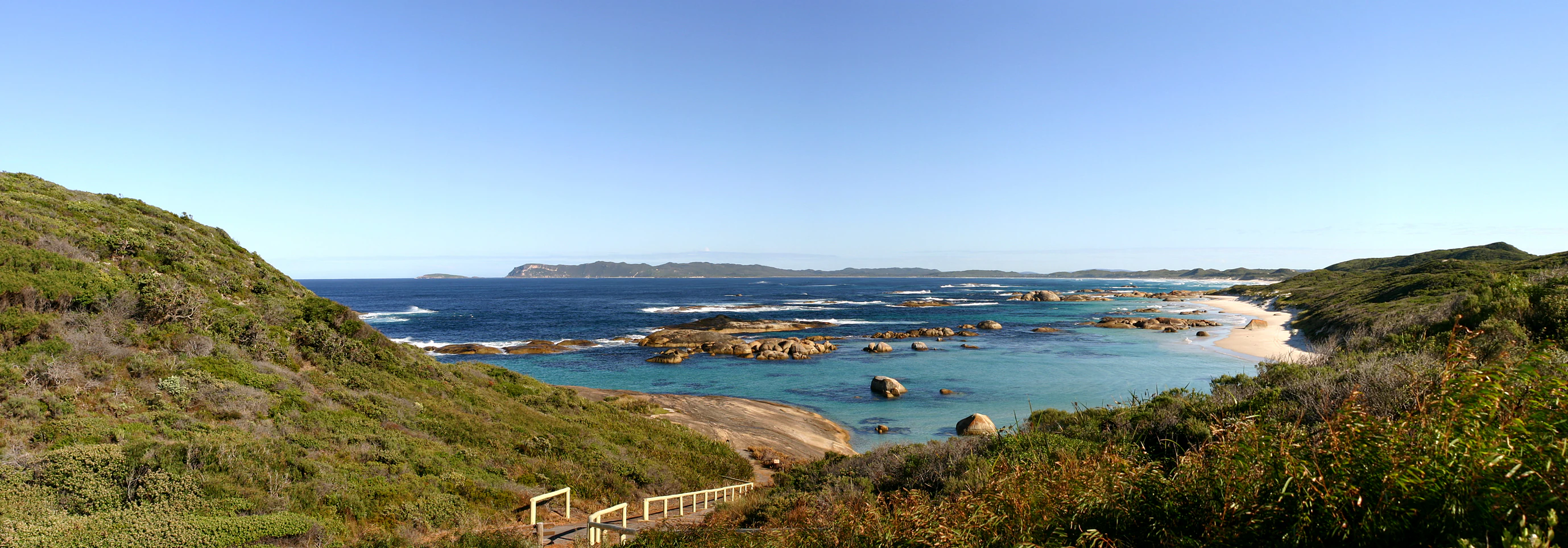 Coastal landscape featuring clear blue water, rocky outcrops, sandy beach, and green hills under a clear sky with wooden railing in foreground.