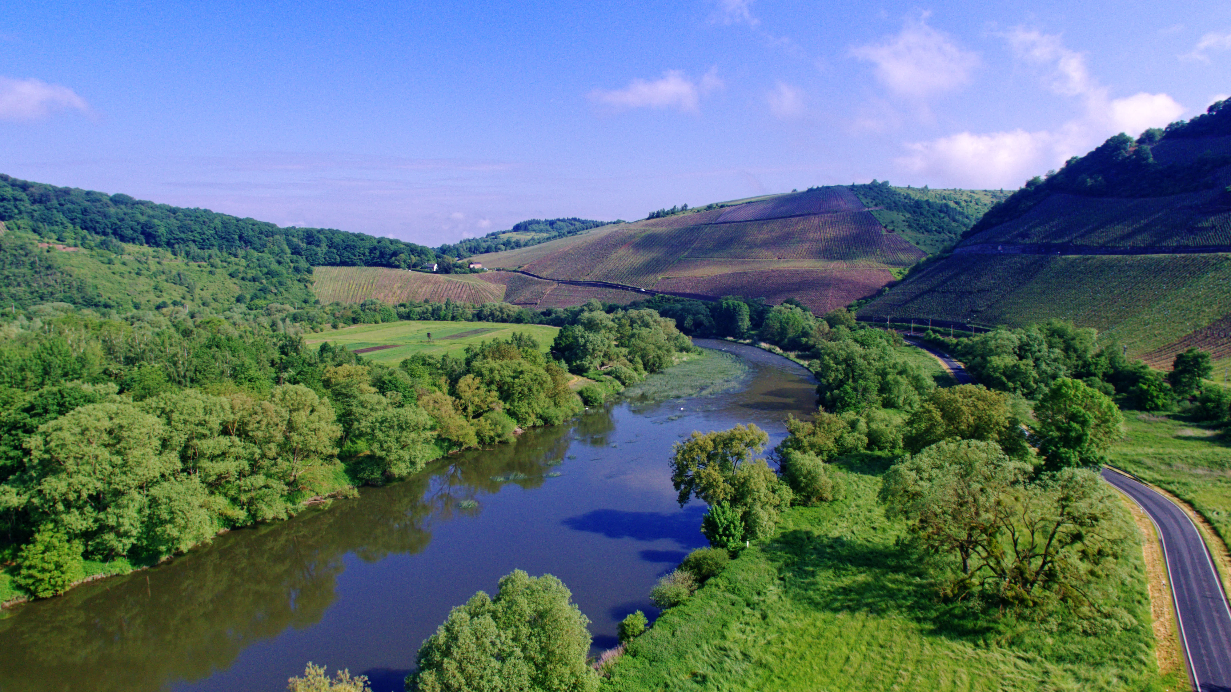 A wide aerial view of the Saar river flowing in a gentle curve through a valley, flanked by lush green trees, fields, and rolling hills.