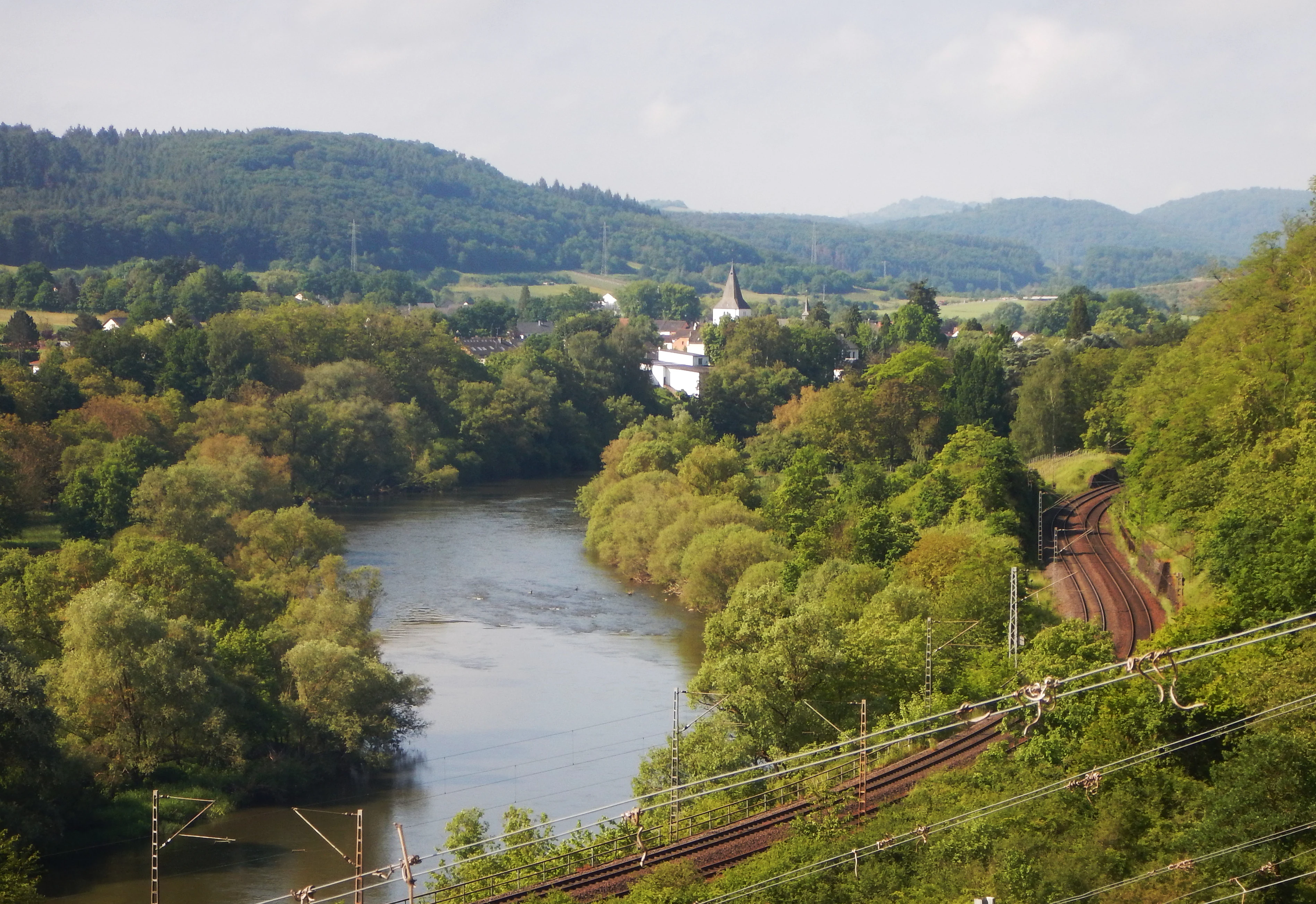 A wide angle view of the Saar river flowing through a forested valley with a railway track visible on the right foreground.