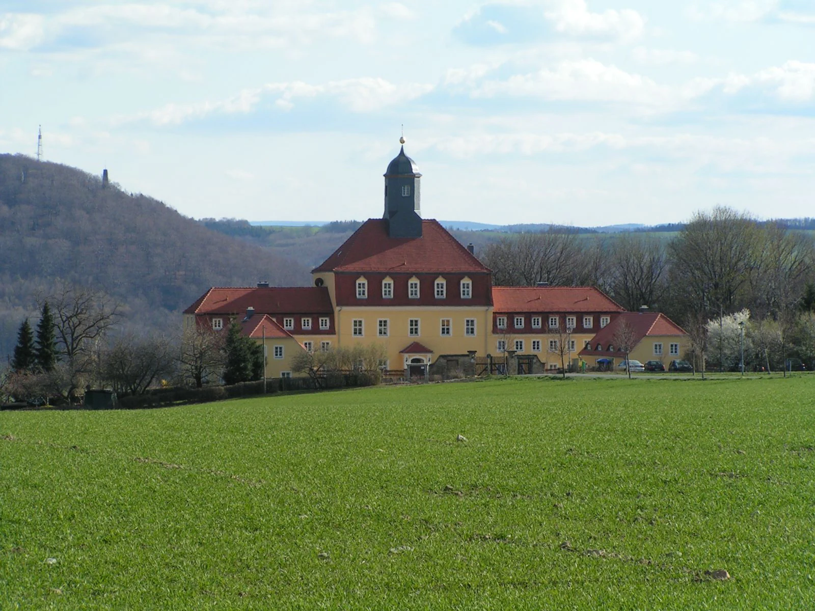A wide landscape view of a yellow two story manor building with a red tile roof and a central tower, set against a green meadow and a forested hill in the background under a blue sky.