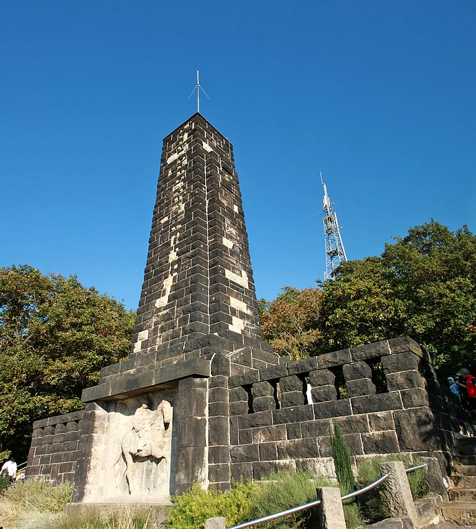 A stone obelisk monument dedicated to King Albert featuring a carved relief panel, set against a bright blue sky and surrounded by dense trees.