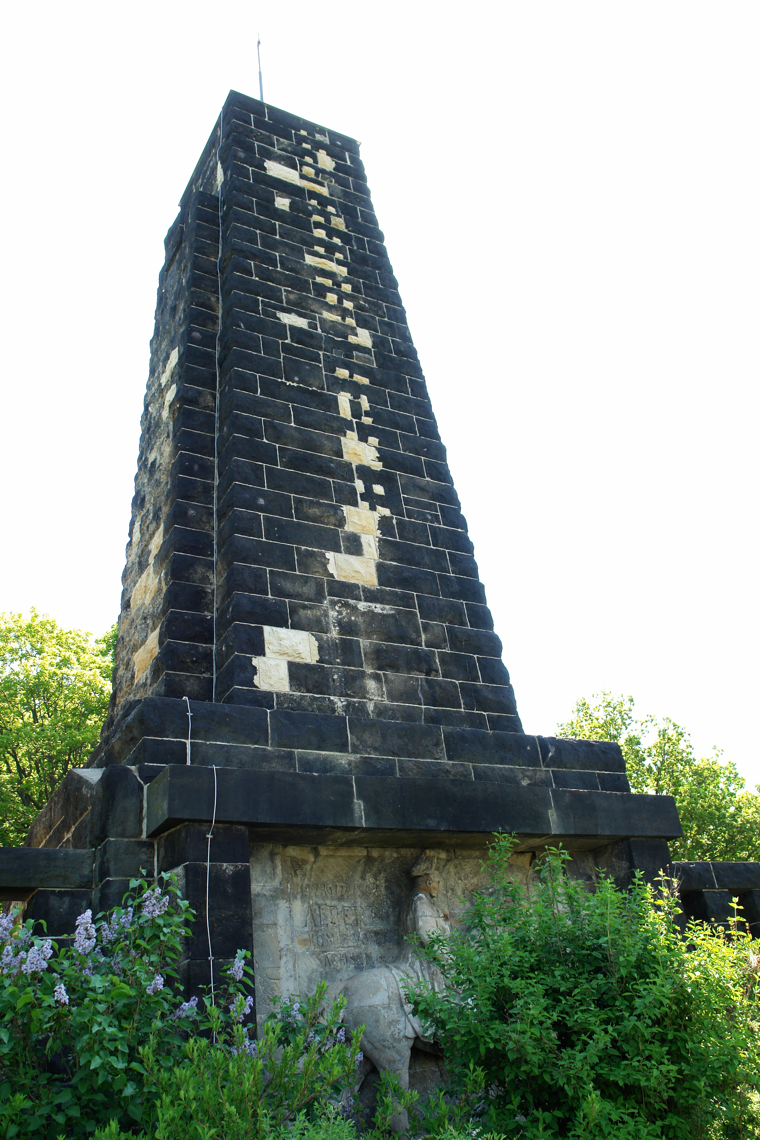 A tall, dark stone monument with a tapered, rectangular pillar shape is partially surrounded by green bushes and trees against a bright, overexposed sky.