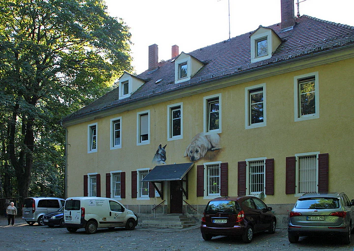 A two story, light yellow building with a gabled roof and brown shutters. It has a small entrance porch and several windows. Several cars are parked in front of the building. Large green trees are visible to the left.