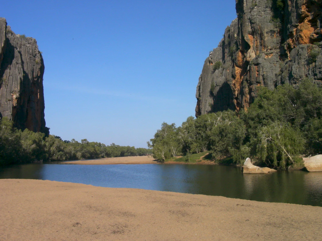 River flowing through a gorge with steep rocky cliffs on both sides, sandy riverbank, and green vegetation along the shore under clear blue sky