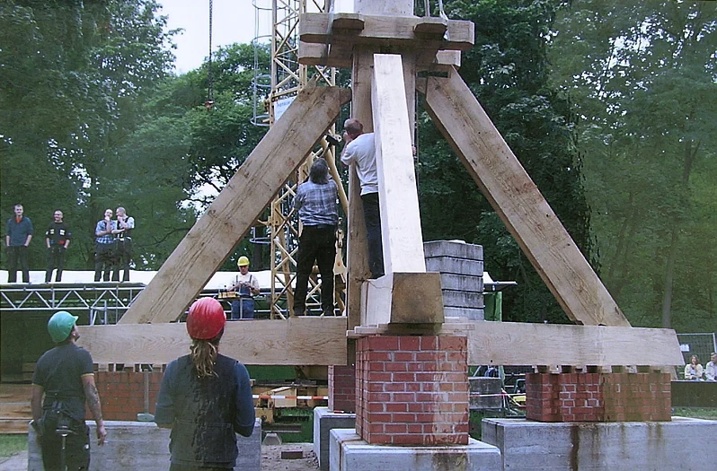 Workers in hard hats assembling the large wooden A-frame trestle of a post mill on brick piers, with scaffolding and onlookers in the background.