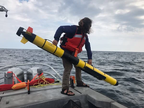 Una persona con chaleco salvavidas está de pie en un barco en el lago Míchigan, sosteniendo un largo vehículo submarino autónomo (AUV) amarillo antes de lanzarlo al agua.
