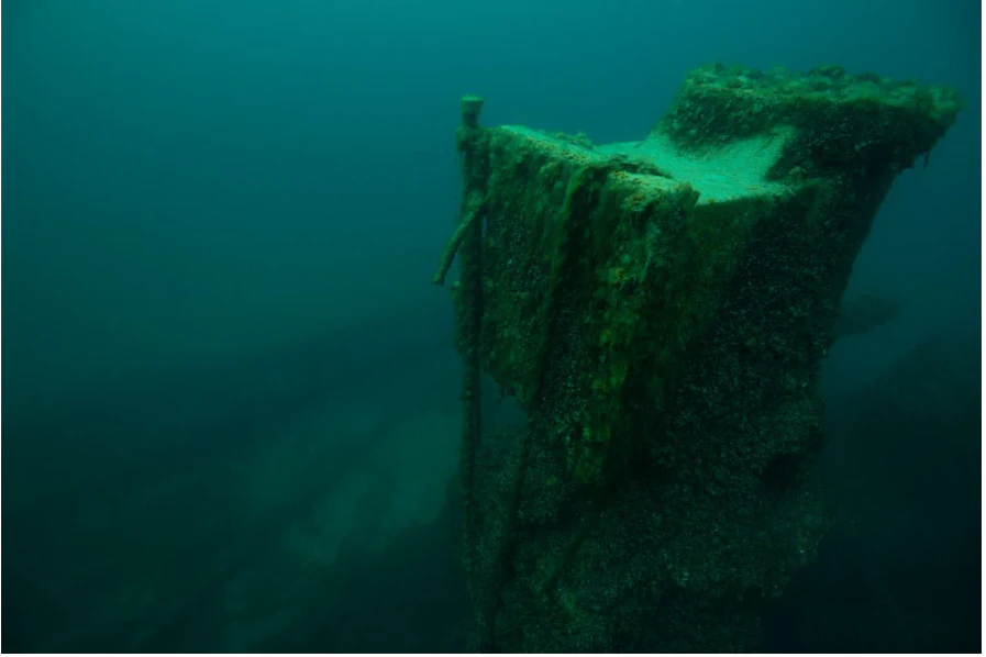 Una fotografía submarina de un mecanismo de máquina de vapor corroído y cubierto de musgo, reposando en el fondo del lago, rodeado de agua oscura, turbia y de un tono azul verdoso.