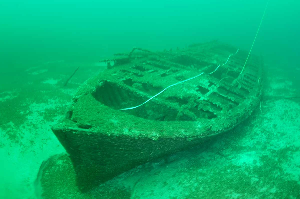 Una vista submarina de los restos de un bergantín de madera naufragado descansando en el fondo arenoso del lago, rodeado de agua verde turbia.