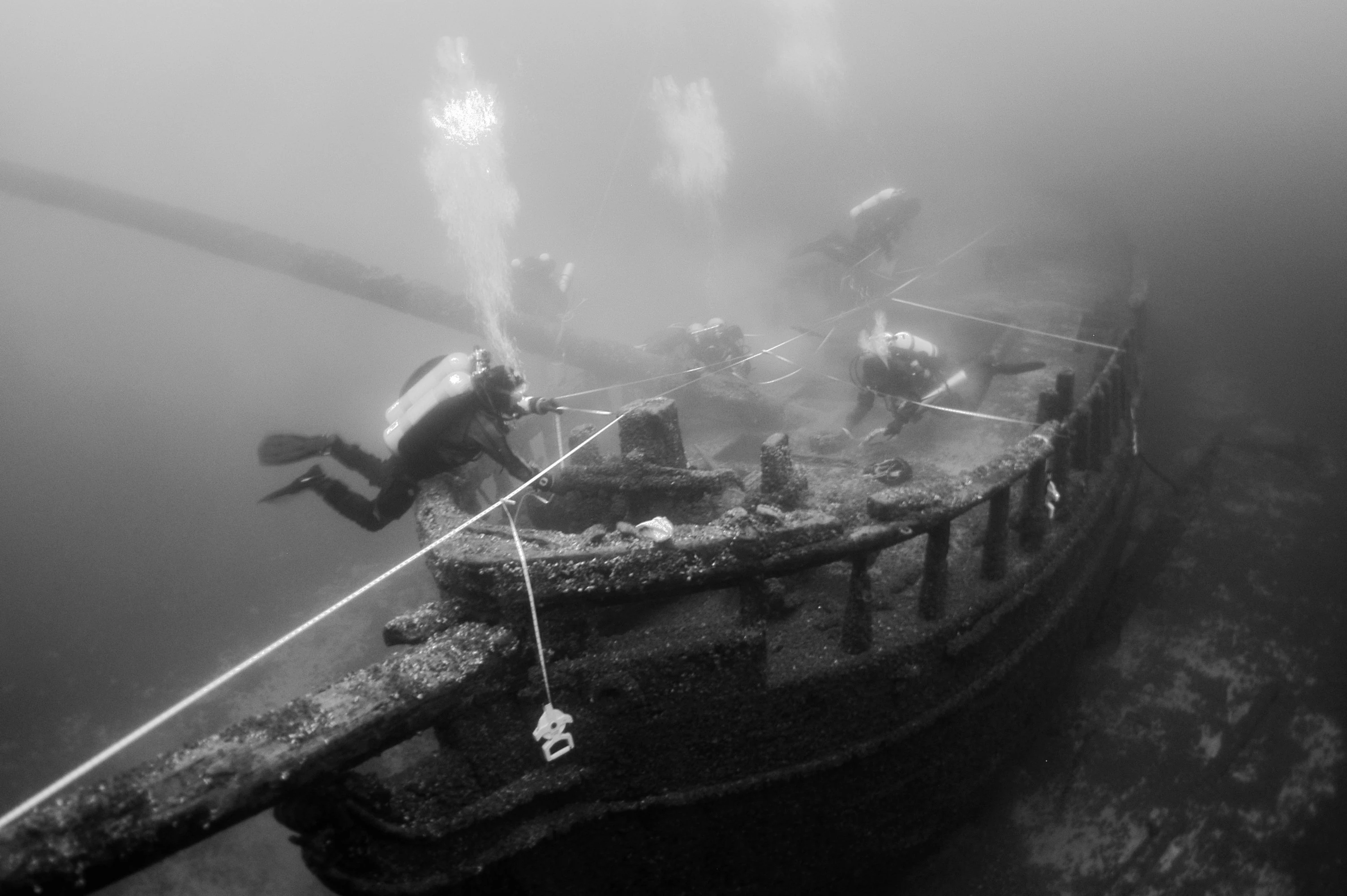Una fotografía submarina en blanco y negro de un naufragio sumergido, con estructuras de cubierta de madera, un mástil y varios buceadores explorando activamente el sitio con líneas de guía.