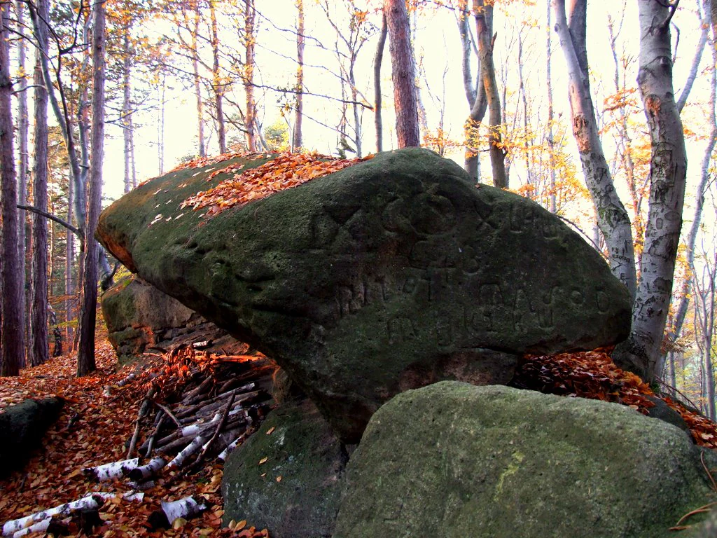 A large, weathered sandstone boulder with inscriptions, situated in a forest with fallen autumn leaves covering the ground.