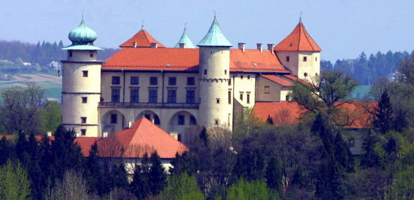 A historic castle with red tiled roofs, multiple towers, and cream colored stone walls, situated amidst a dense green forest canopy.