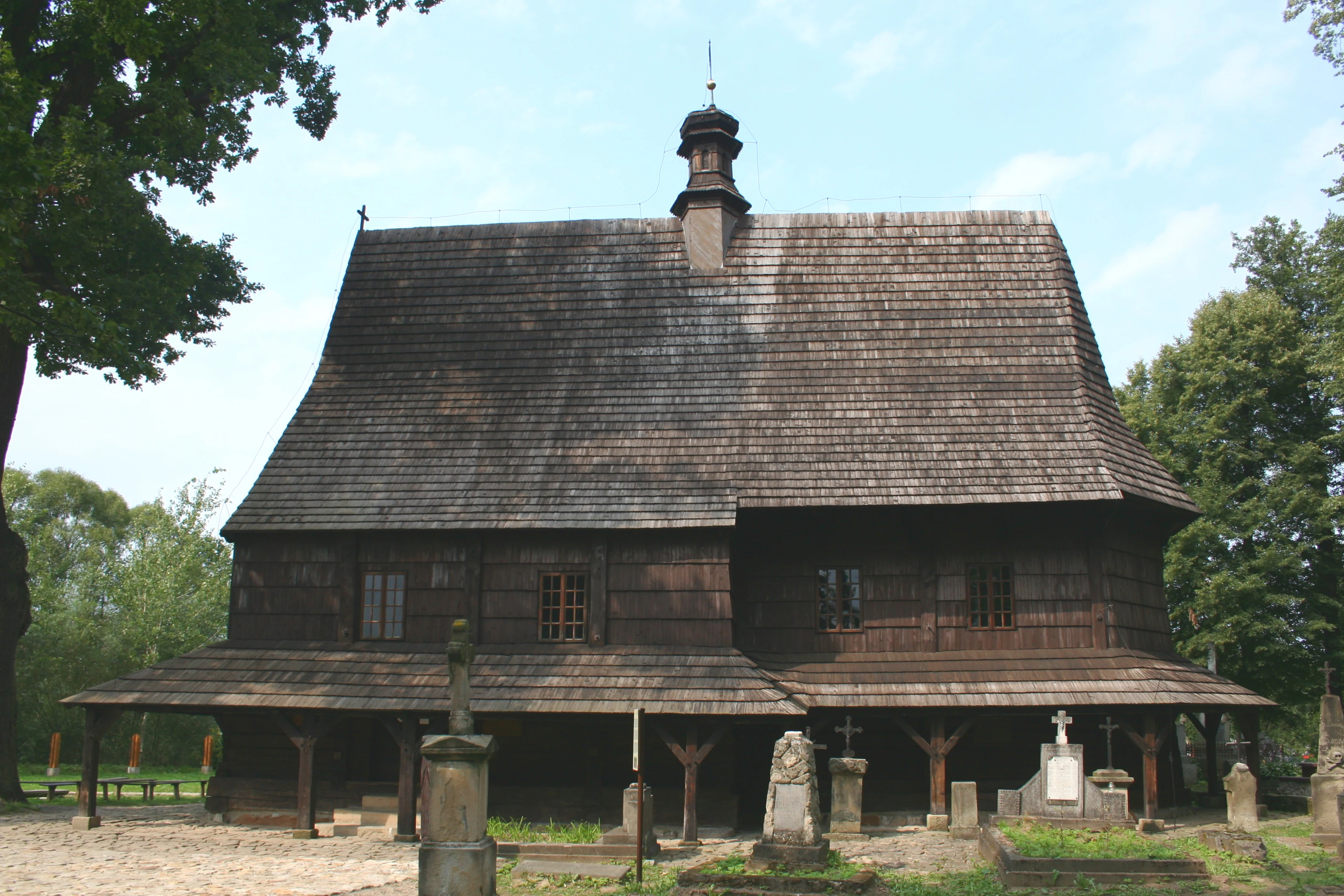 Front view of the historic wooden Saint Leonard Church in Lipnica Murowana, featuring a steep shingled roof, dark timber walls, and surrounding stone grave markers in the foreground.