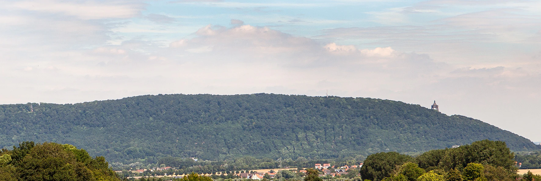 A wide shot of a forested hill ridge under a cloudy sky, with a small structure visible on the crest of the hill.