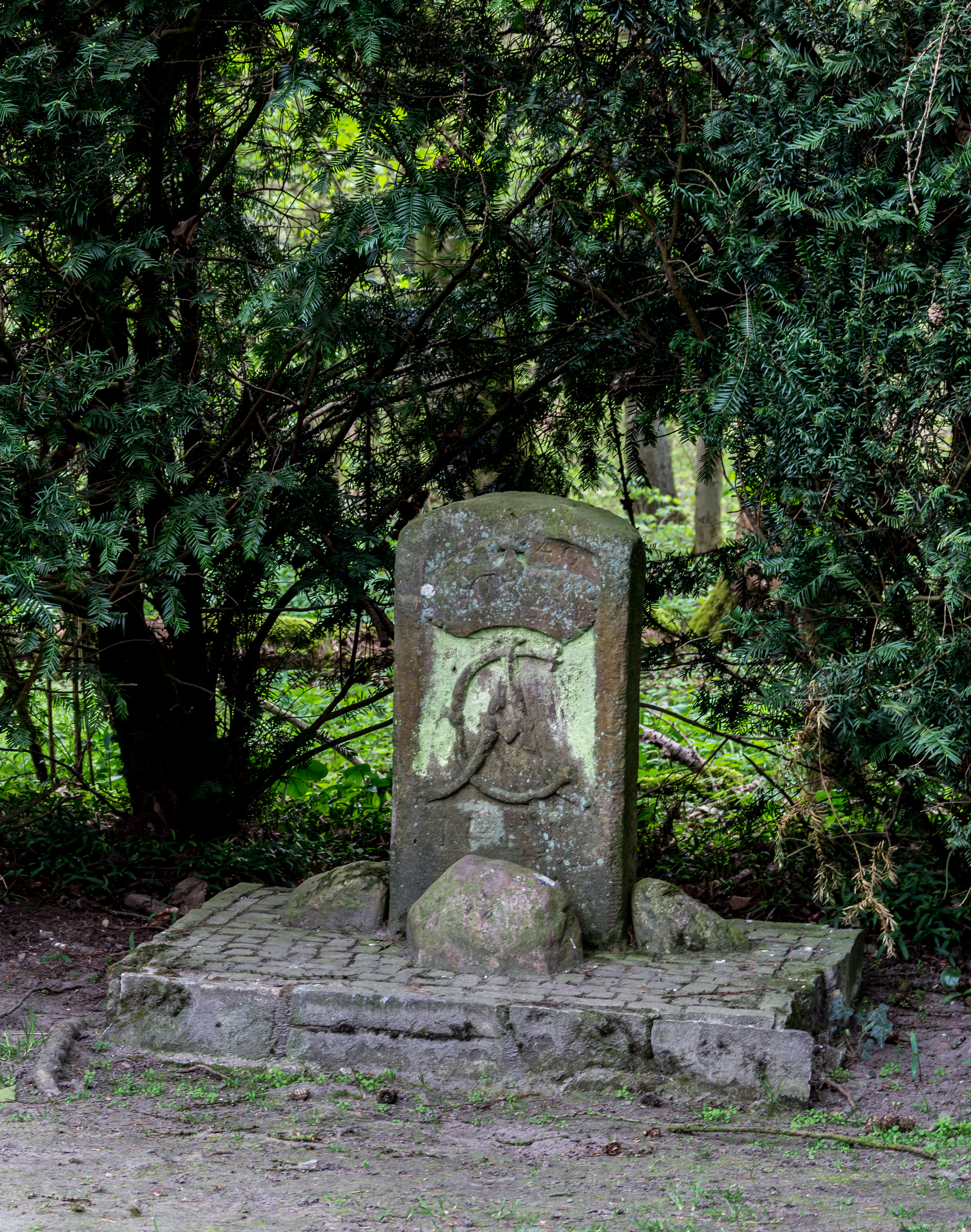 A weathered stone monument stands on a small paved stone platform in a wooded area, featuring an carved crest and initials.