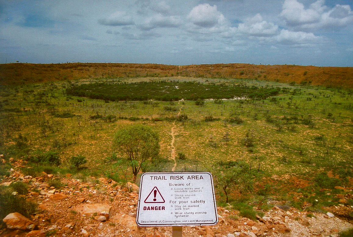 Warning sign reading "Trail Risk Area" in foreground with crater visible in background across grassy and rocky landscape under partly cloudy sky