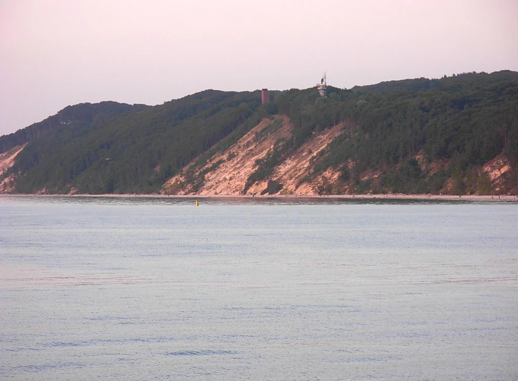 Calm sea with sandy and forested cliffside featuring a small structure on the hilltop