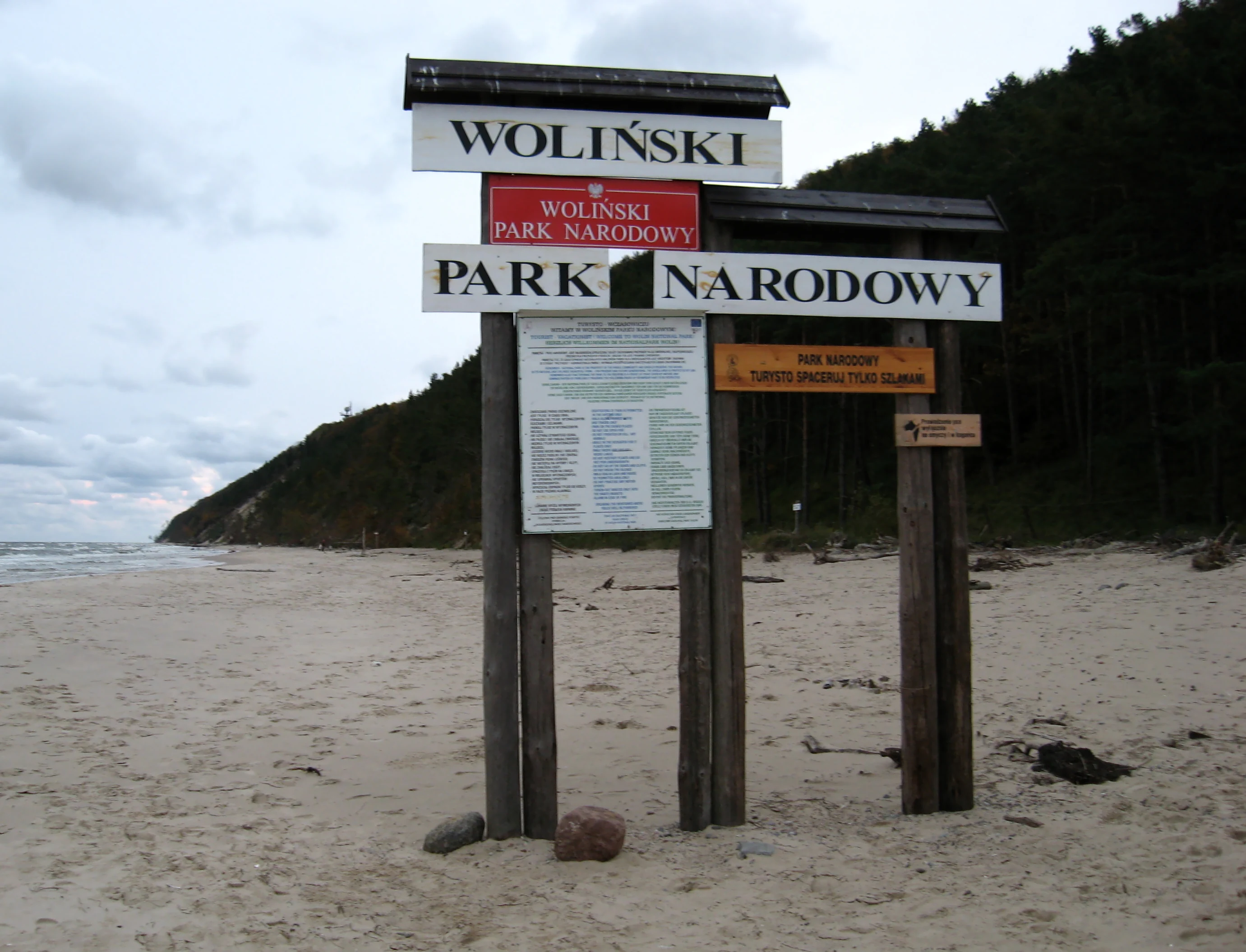 A multi-tiered wooden park sign displaying the text Woliński Park Narodowy, standing on a sandy beach with a forest-covered cliff and the sea visible in the background.
