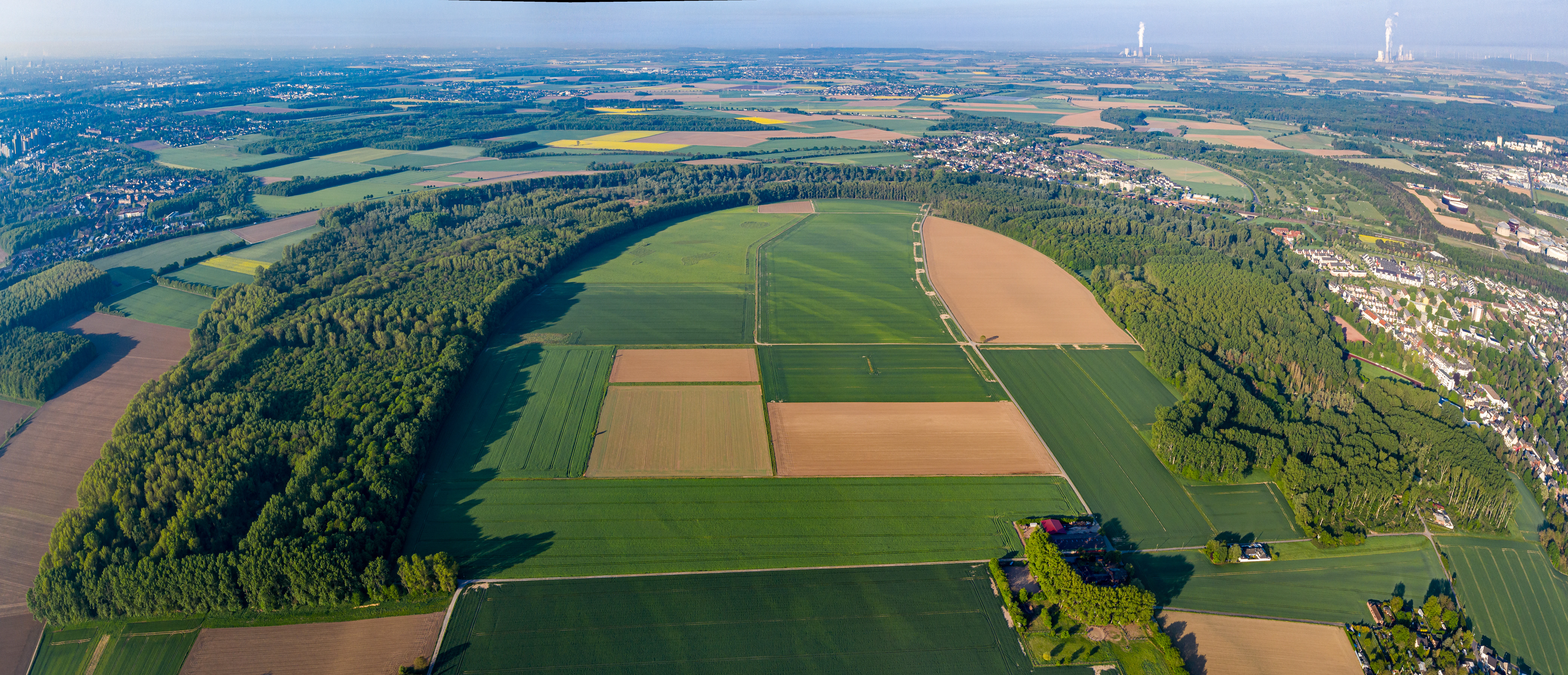A wide aerial panoramic shot showing a dense, curved band of forest surrounding a mix of agricultural fields near a suburban area.