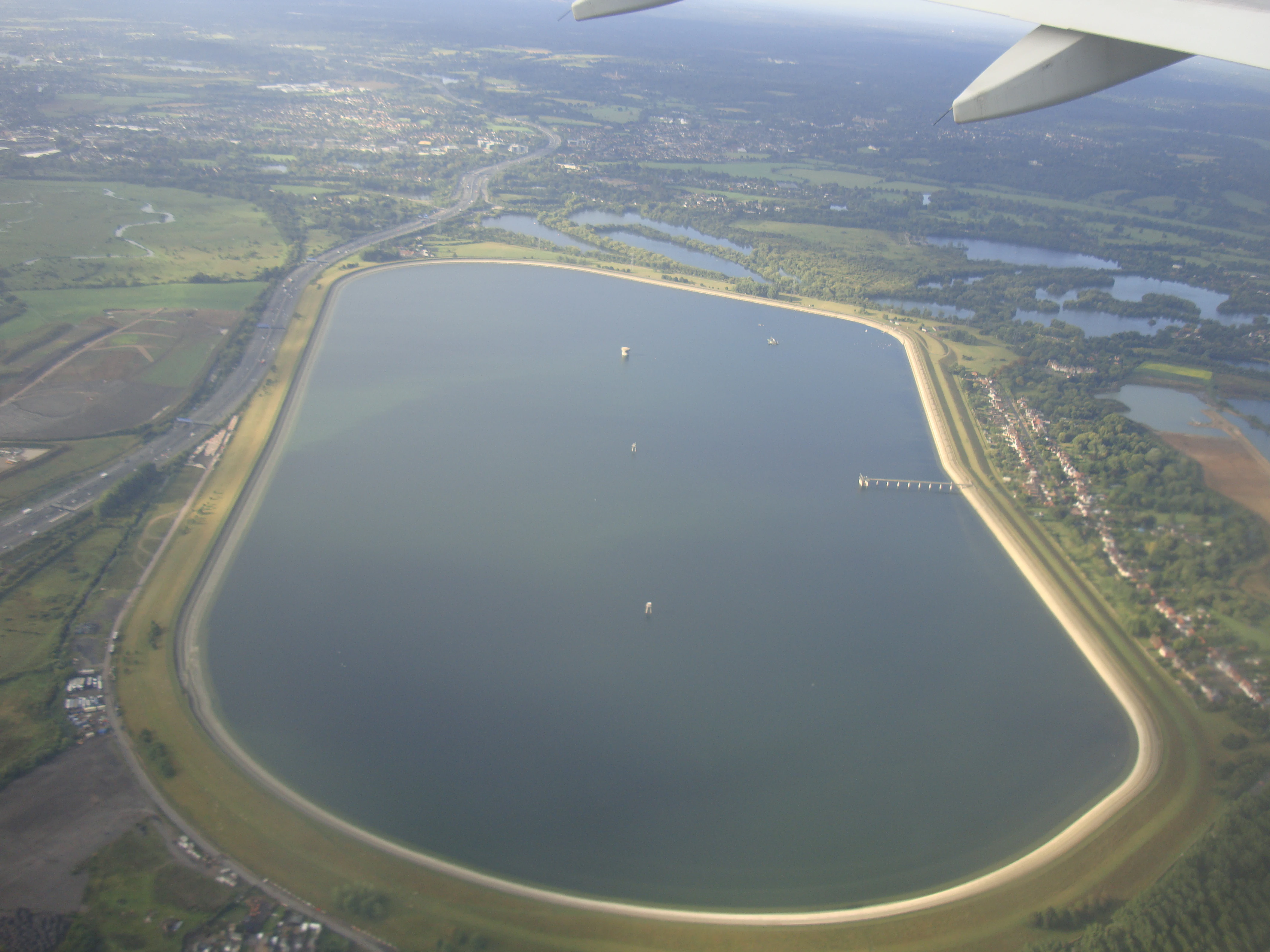 Aerial view looking down at the oval-shaped Wraysbury Reservoir, surrounded by green fields and nearby roads, with the wing of an aircraft visible in the top right corner.