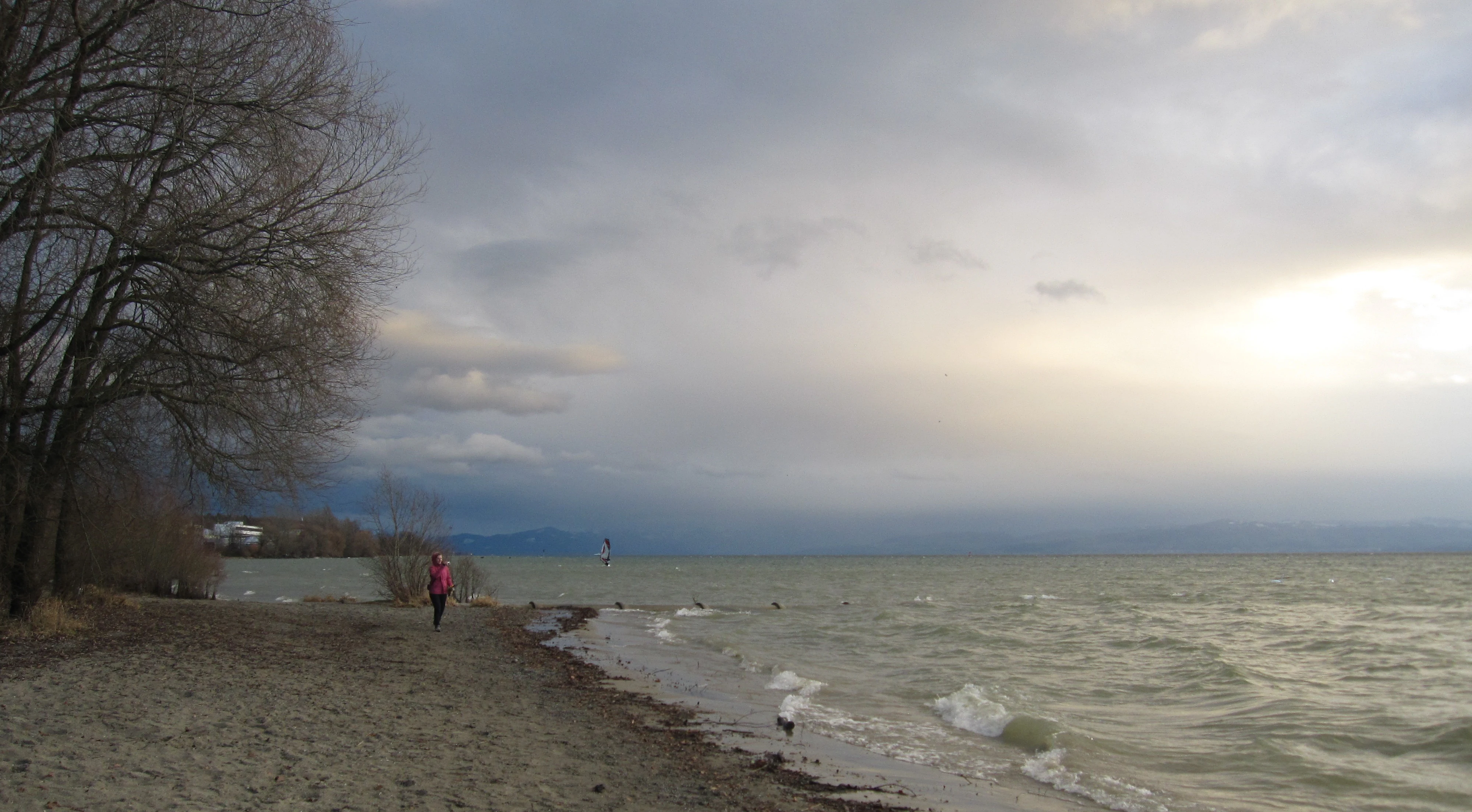 A person walking on a sandy beach along the shoreline of a large, wavy lake under a cloudy sky.
