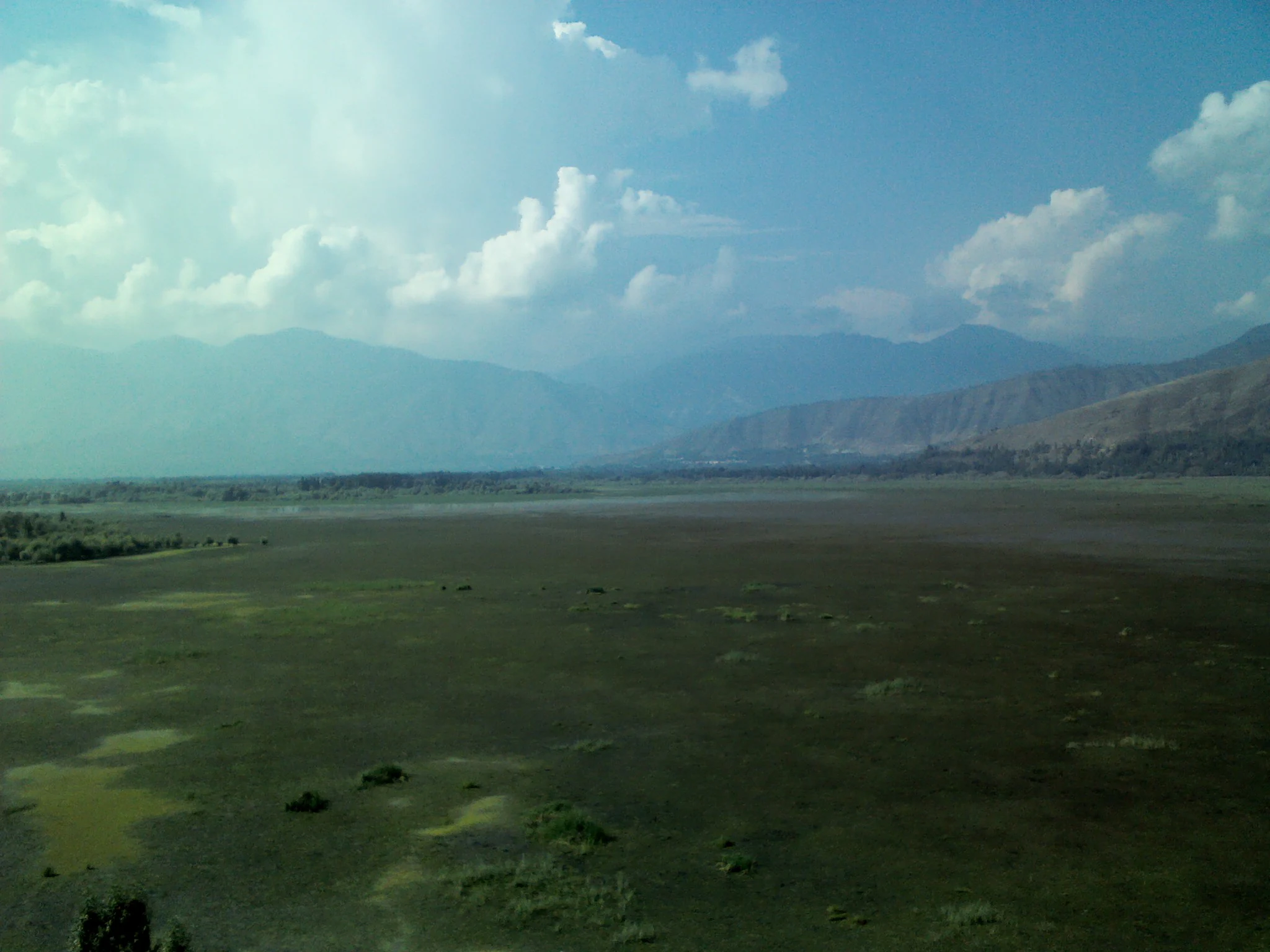 A wide, low-angle landscape view looking across a large, flat, marshy expanse toward distant rolling mountains under a blue sky with scattered clouds.