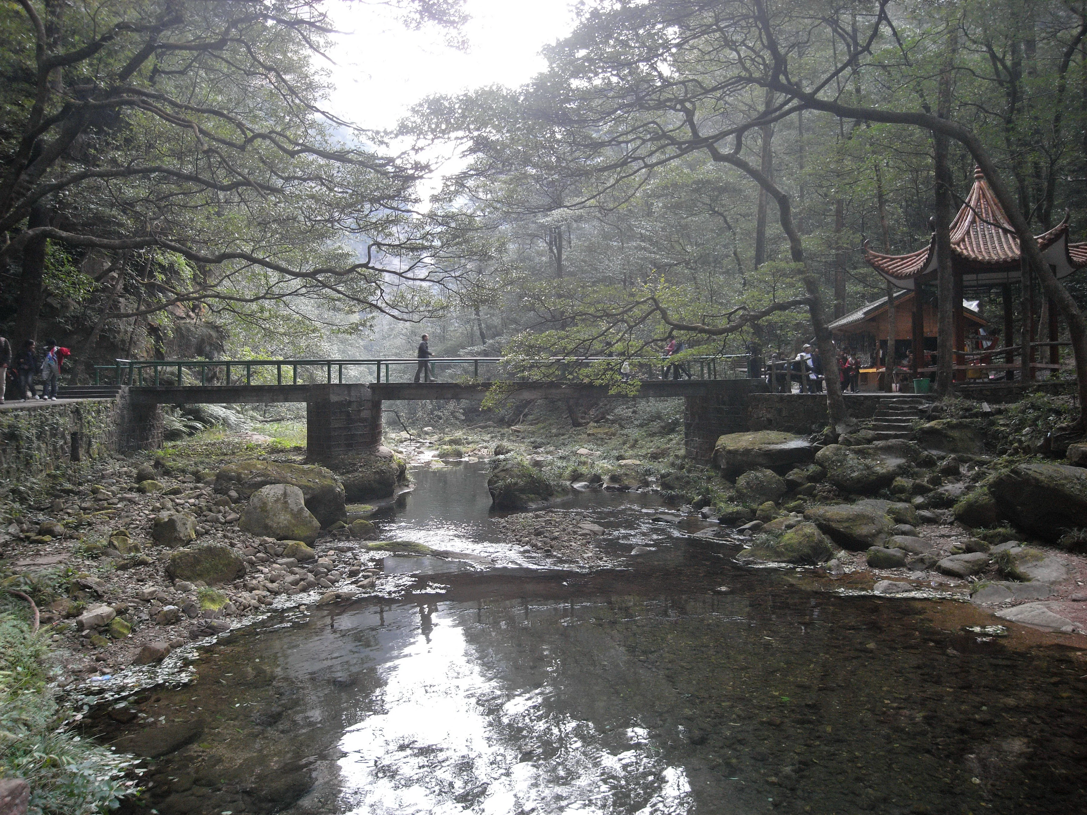 A stone footbridge spans a clear stream in a lush, wooded valley, with a traditional Chinese-style pavilion visible on the bank.