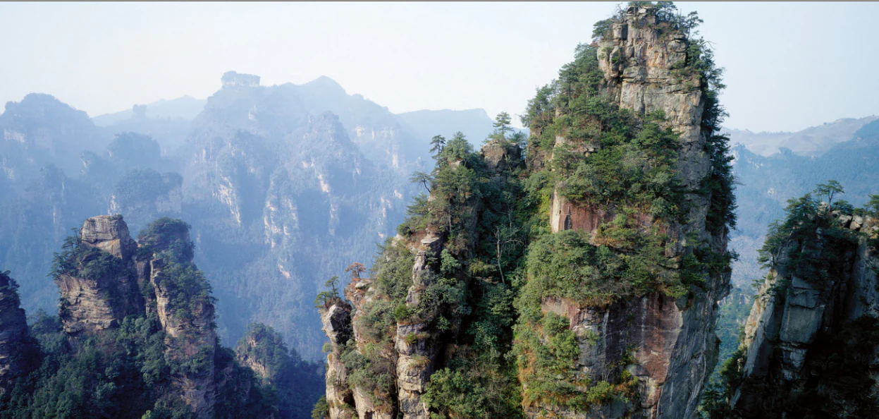 A wide, hazy mountain landscape in China featuring multiple vertical, pillar-like sandstone rock formations covered in green vegetation.