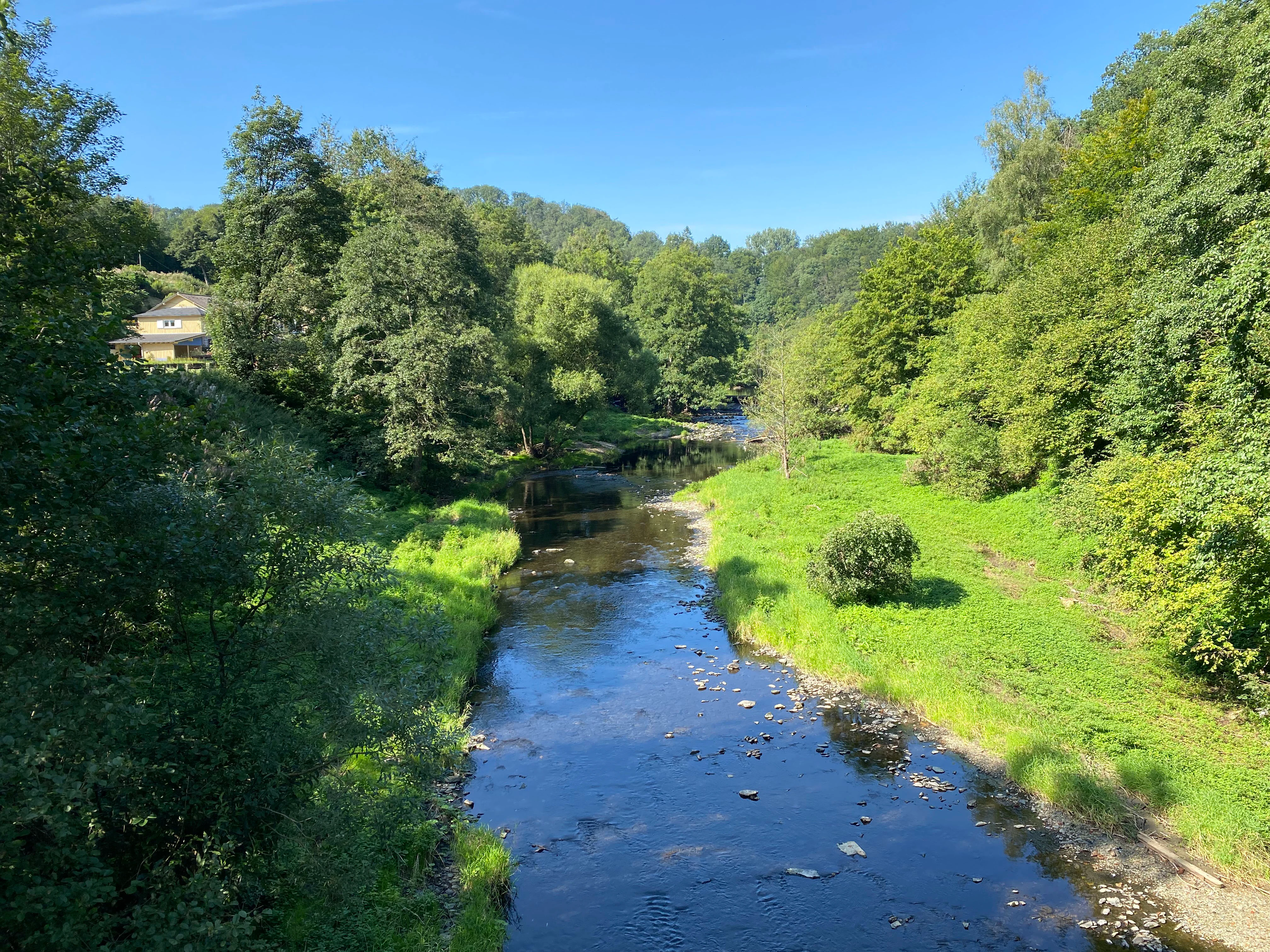 A calm, narrow river flows through a vibrant green valley, flanked by dense trees and shrubs under a clear blue sky, with a small house visible on the left bank.