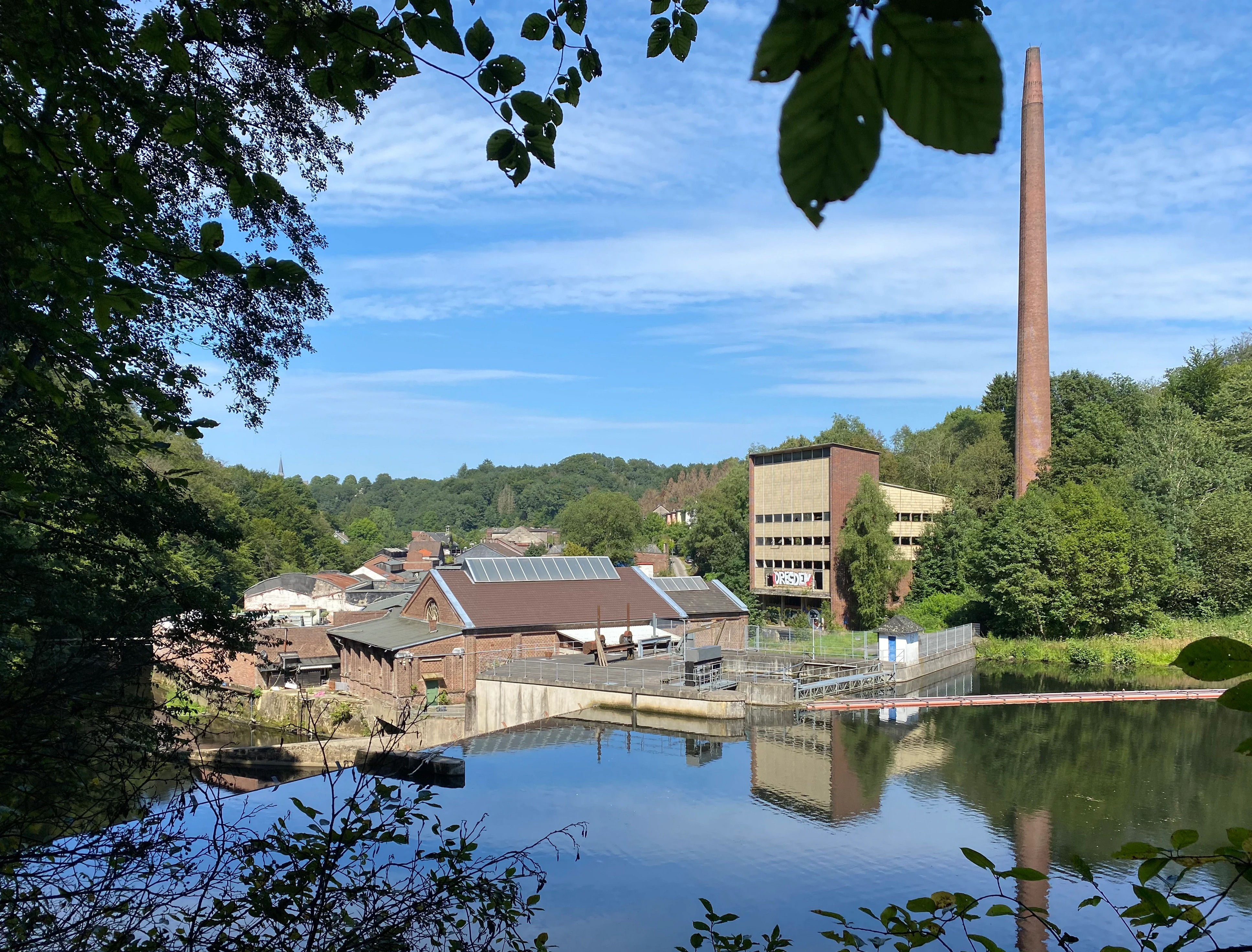 A view of the industrial Stauanlage Dahlhausen dam and brick building on the Wupper river, framed by trees in the foreground, with a tall industrial chimney visible under a bright sky.