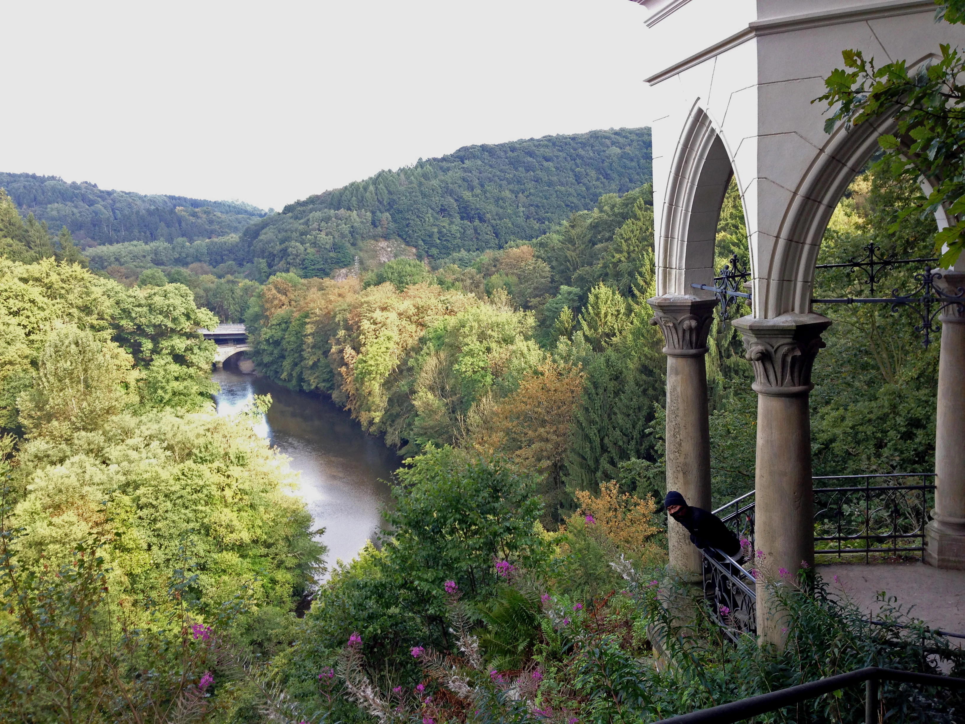 A stone pavilion with arched pillars overlooks a forested valley and a river flowing through the center, with a small bridge visible in the distance.