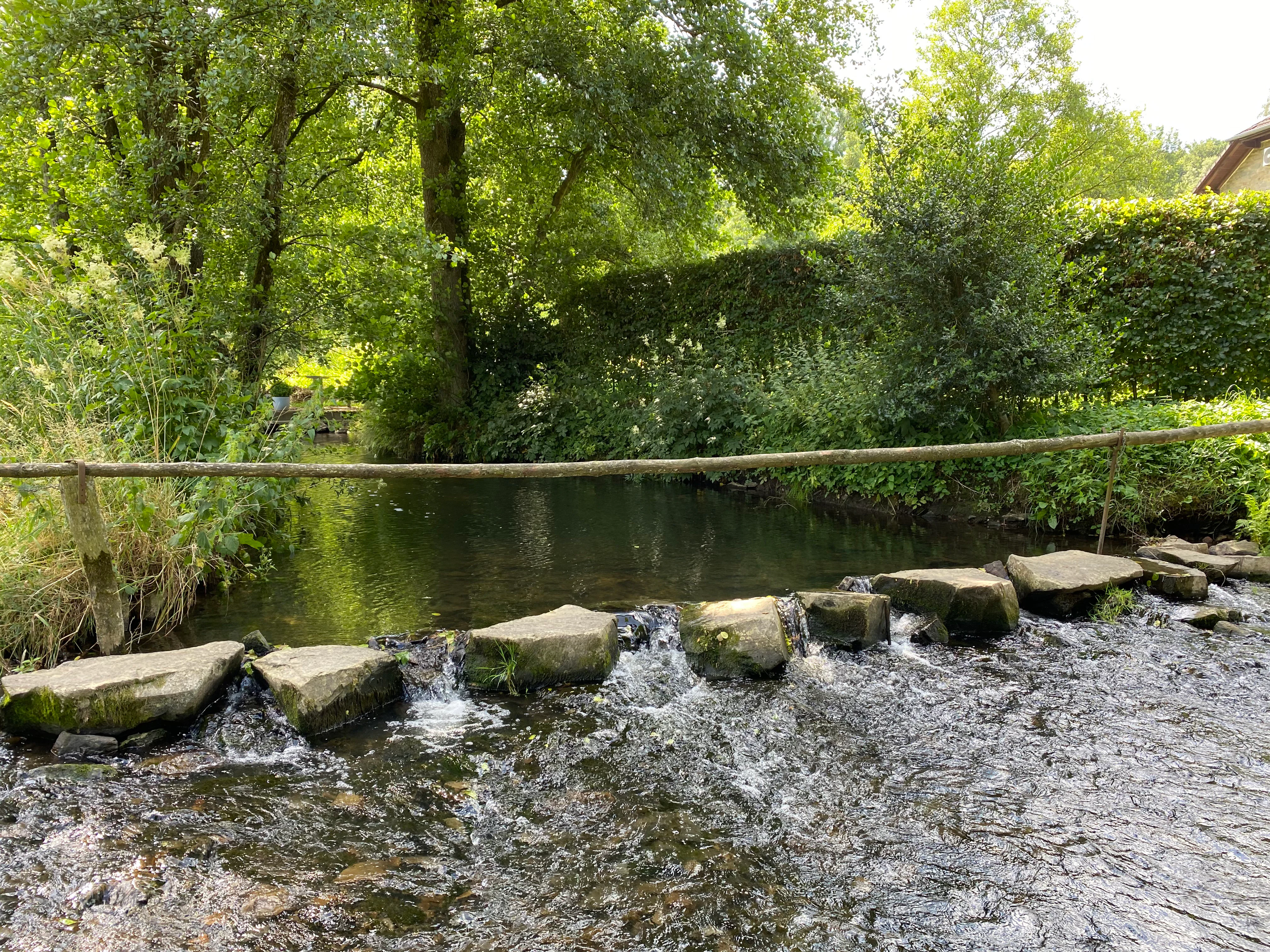 A shallow river ford with flat stone stepping stones crossing the water, bordered by dense green trees and a wooden rail fence.