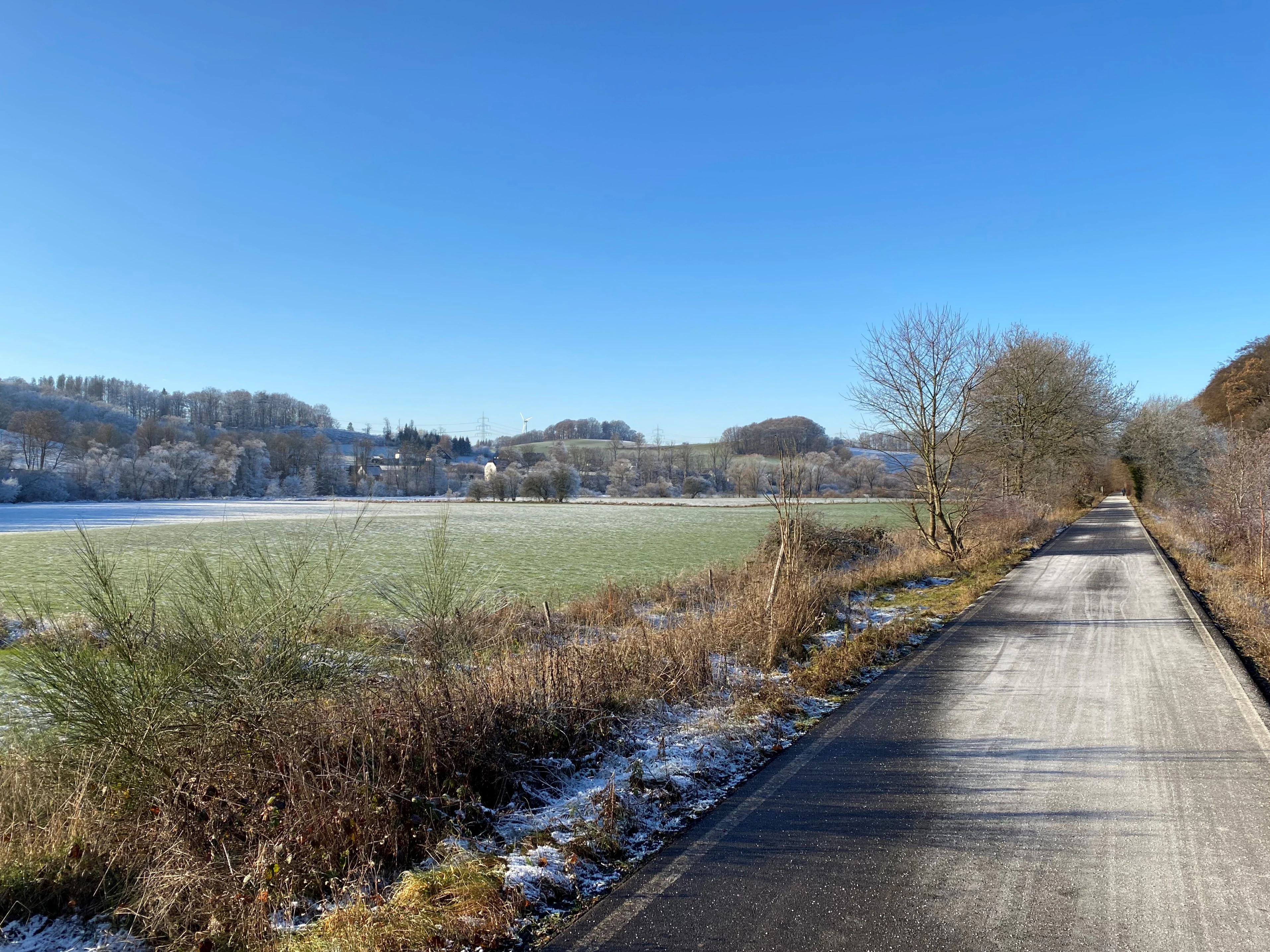 A paved bicycle path stretches along the side of a field covered in a light dusting of frost, with a clear blue sky above and distant hills in the background.
