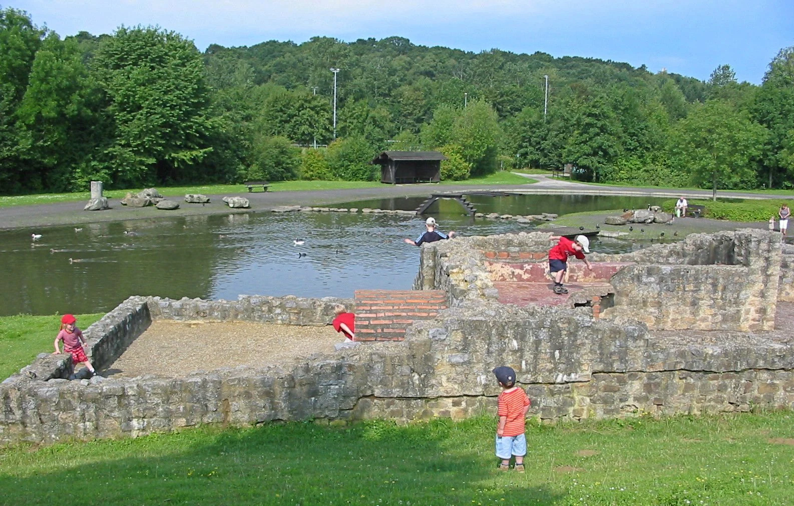 Children playing near the low stone walls of the reconstructed Roman bath ruins, set against a small pond and green forested hillside.