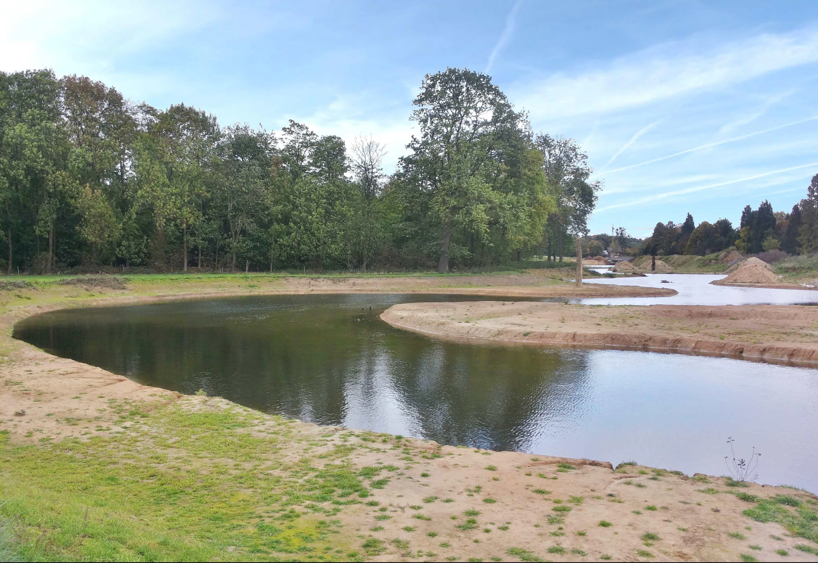 A newly realigned, winding river channel with exposed sandy banks, bordered by green trees and fields on a clear day.