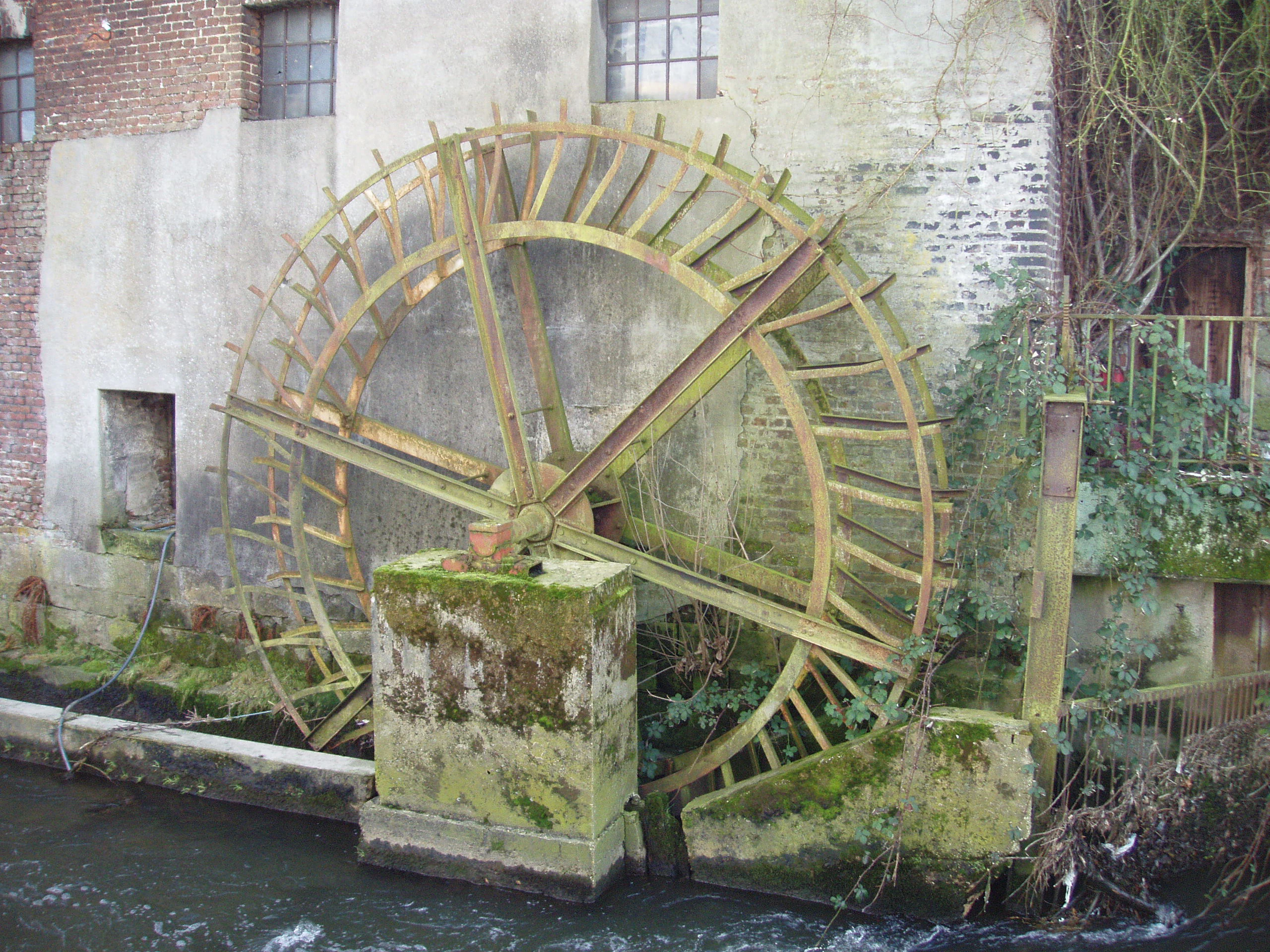 A close up view of a large rusted metal watermill wheel partially submerged in a flowing river next to a stone and brick building wall.