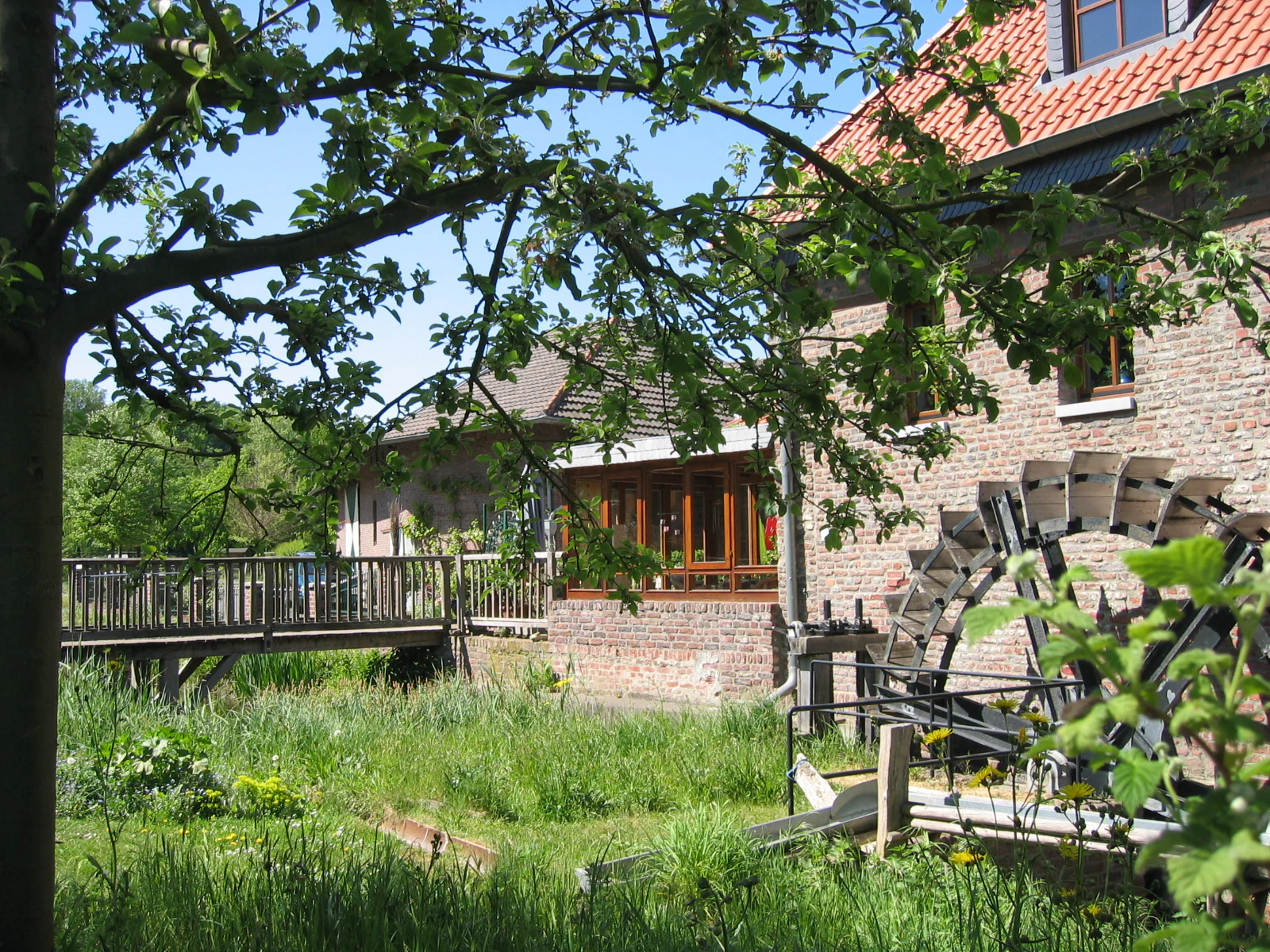 A historic brick building with a water wheel in the foreground, situated in a green, grassy valley area with a wooden bridge to the left.