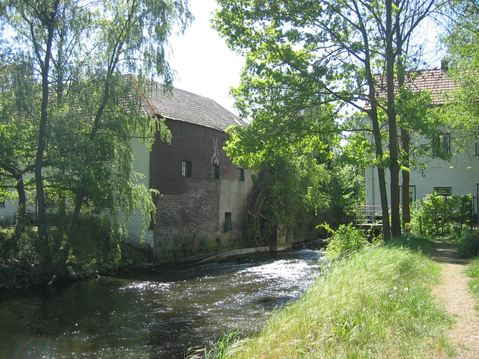 An old brick and stone mill with a water wheel located on the edge of a flowing river, surrounded by lush green trees and a dirt path.