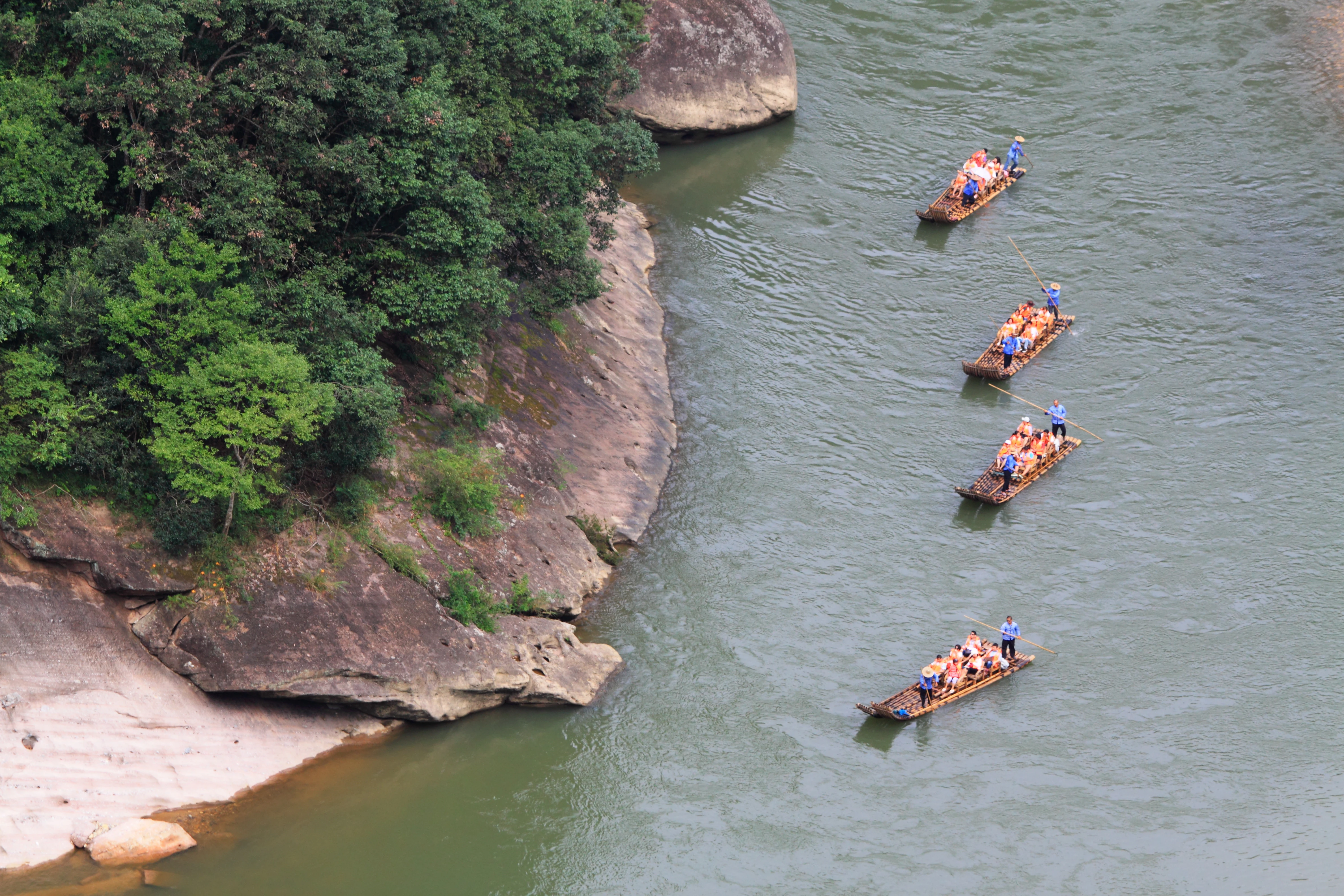 A high-angle view looking down at a river winding past rocky, tree-covered cliffs, with four bamboo rafts carrying tourists floating downstream.