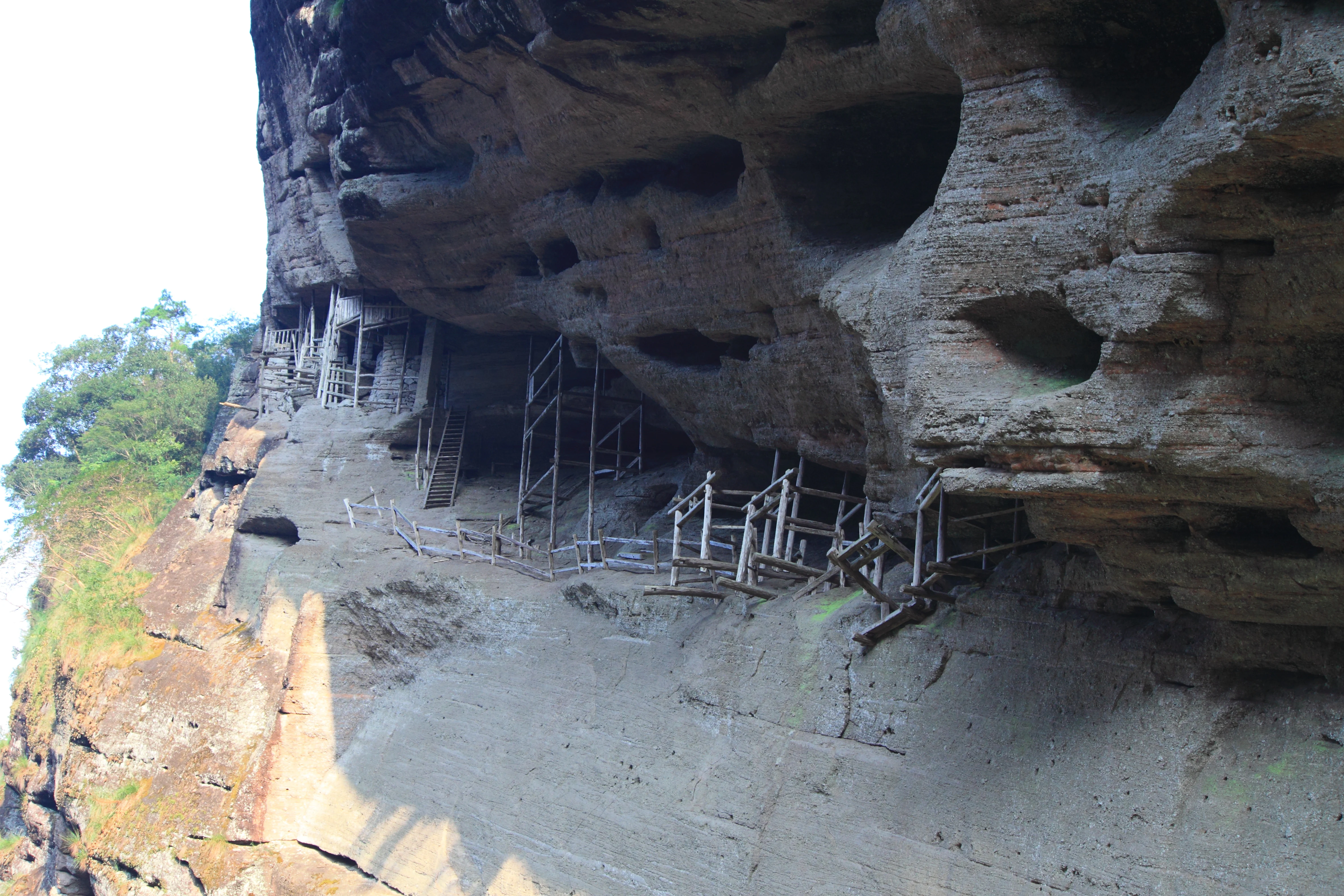 Ancient dwellings tucked into a large, rocky cliffside cave overhang in the Wuyishan Scenic Area, featuring wooden support scaffolding.