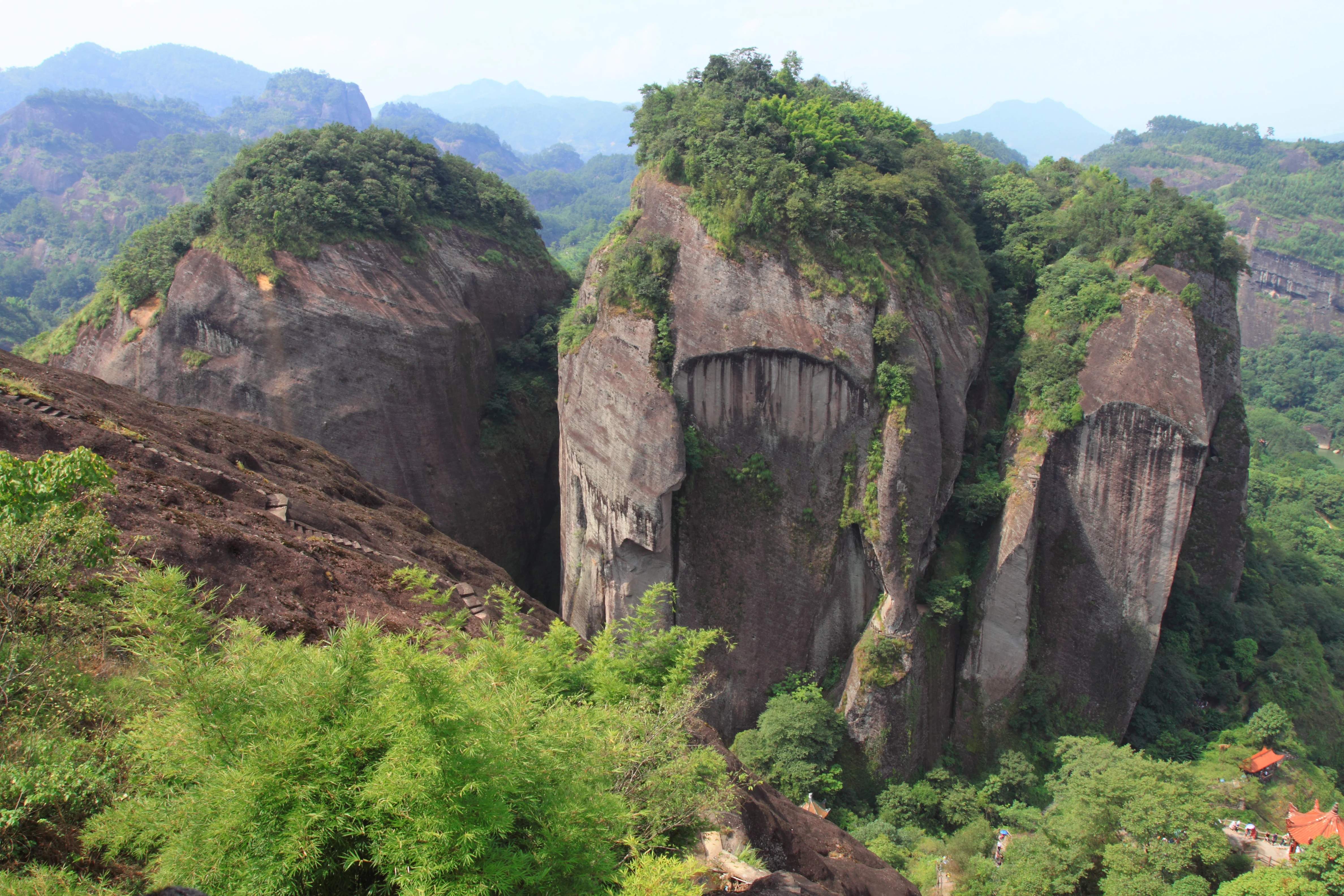 A wide angle view of massive, steep rocky cliffs covered in patches of forest, characteristic of the Danxia landforms in Wuyishan Scenic and Historic Interest Area.