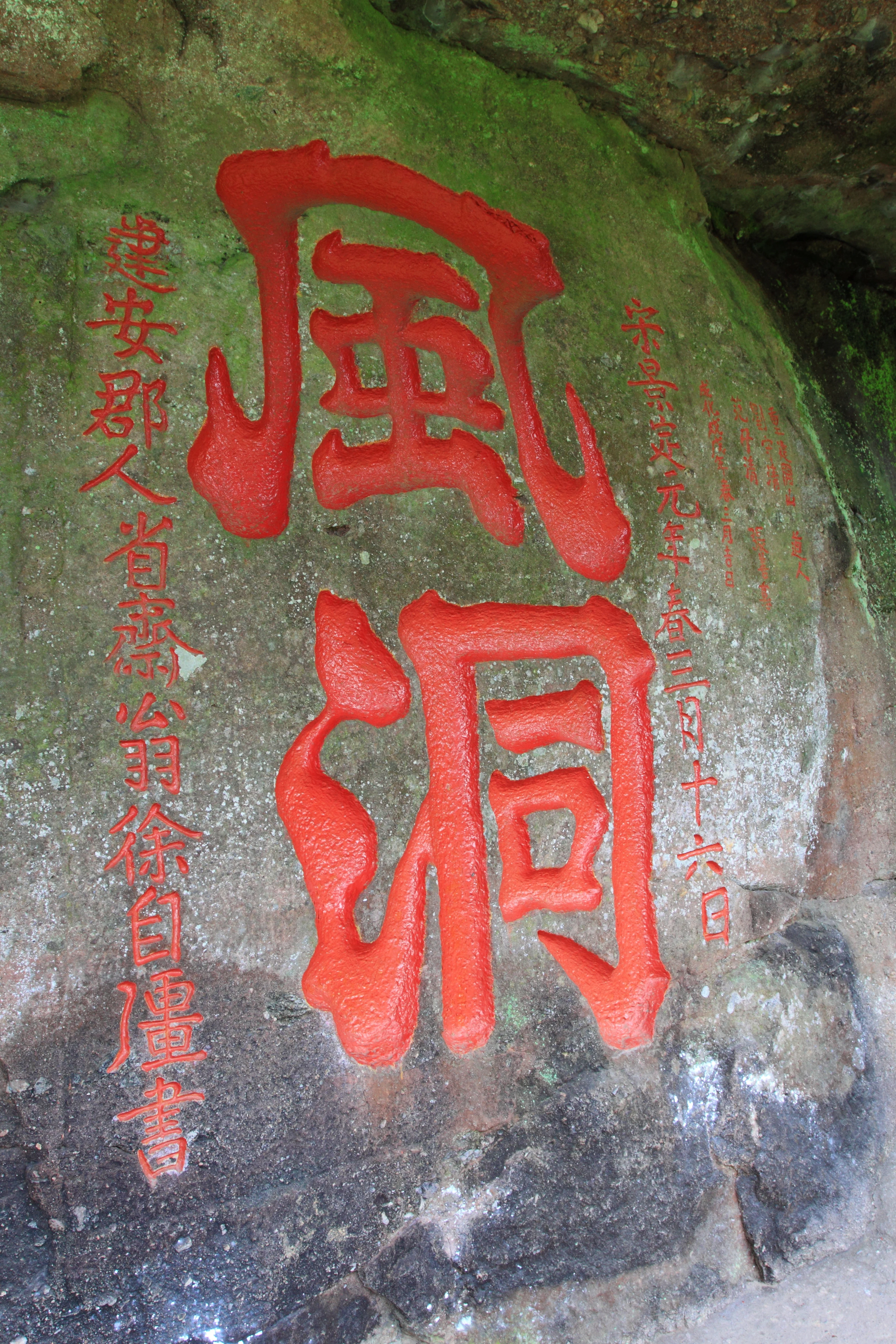 A close-up view of red Chinese calligraphy carved into a natural mossy rock face.