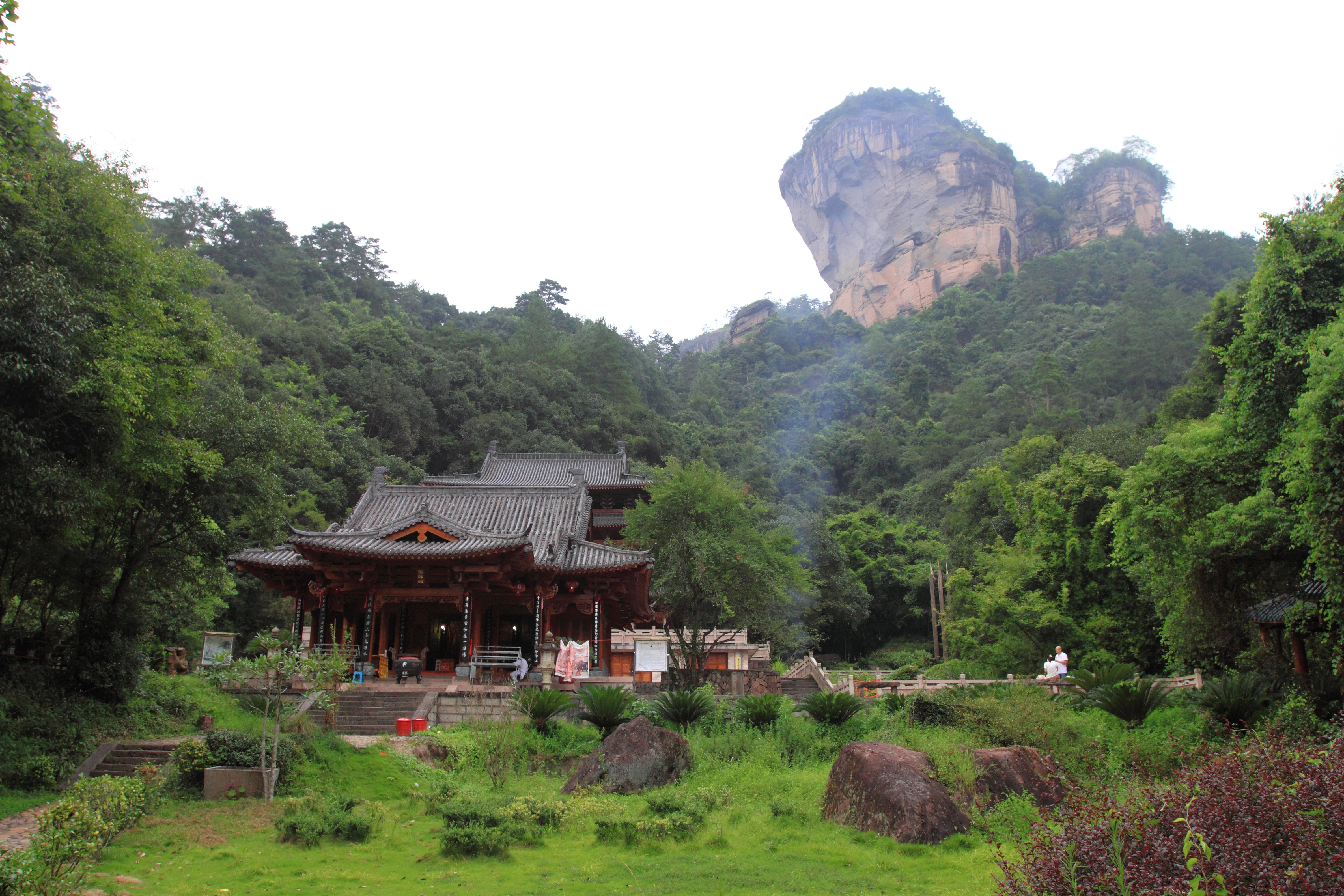 A traditional Chinese temple building with dark tiled roofs sits at the base of a lush, forest-covered hill, with a large, sheer granite-colored peak rising prominently in the background under a hazy sky.