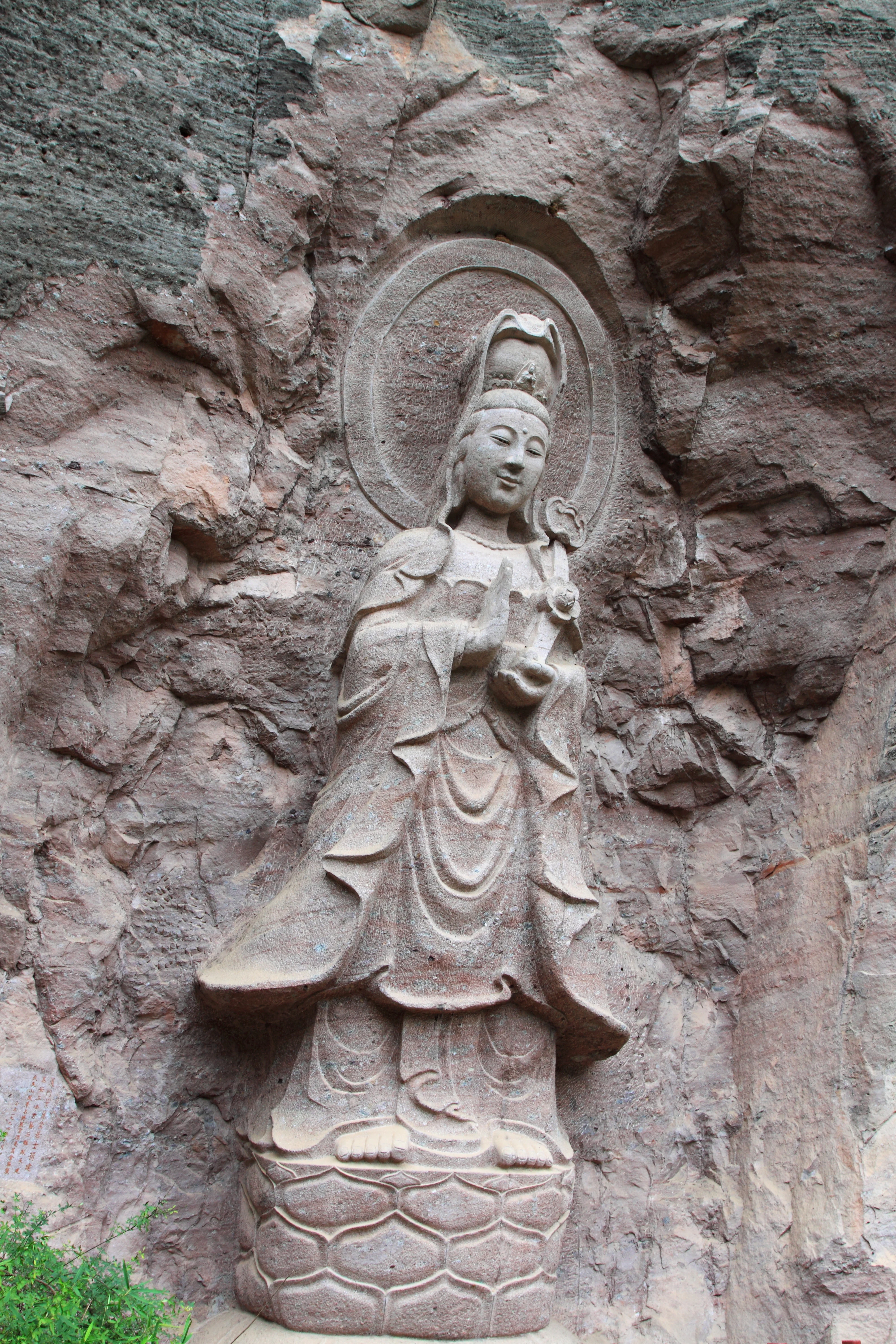 A carved stone statue of the Buddhist figure Guanyin standing on a lotus base, integrated into a rough, reddish-brown rock cliff face.